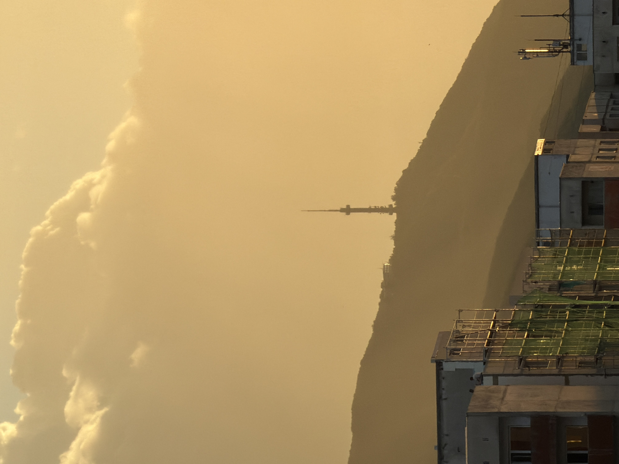 The image depicts a cityscape at dusk, likely in Sofia, Bulgaria, with a prominent mountain range in the background. The sky is filled with a warm, golden hue, characteristic of sunset, with soft, diffused clouds in the upper portion.
In the foreground, several multi-story buildings are visible. One building on the left appears to be under construction, with scaffolding and green netting covering its facade. Two individuals are seen standing on the roof of another building on the left, seemingly observing the surroundings. The architecture of these buildings suggests a residential or mixed-use urban environment.
Further in the background, the silhouette of a mountain is discernible, with a radio tower perched atop its peak. The mountain is bathed in the soft, fading light, creating a sense of depth and scale. The overall atmosphere is tranquil and serene, capturing the quiet beauty of the city as day transitions into night.