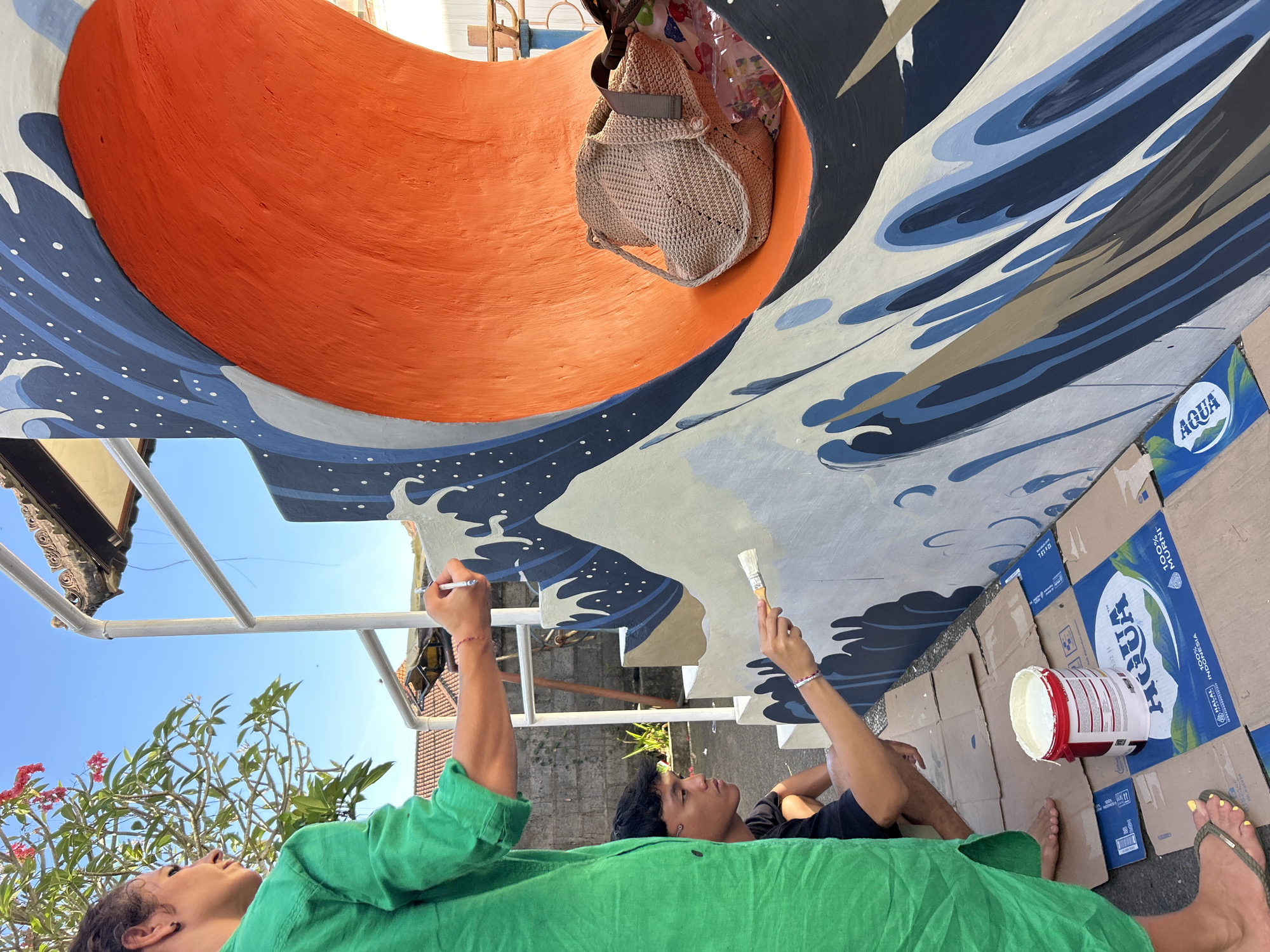 The image shows two individuals painting a mural of ocean waves and a circular orange shape on a wall in Kediri, Indonesia.
One person, wearing a green shirt, is lying on the ground, looking up at the mural and holding a paintbrush. The second person, wearing a black shirt, is lying next to them, also looking up and holding a different paintbrush. The paint is white.
The mural is primarily blue, white, and orange. Cardboard boxes, some marked with "AQUA" branding, are spread on the ground beside them.
The setting appears to be outdoors, with visible vegetation, buildings, and a blue sky.
