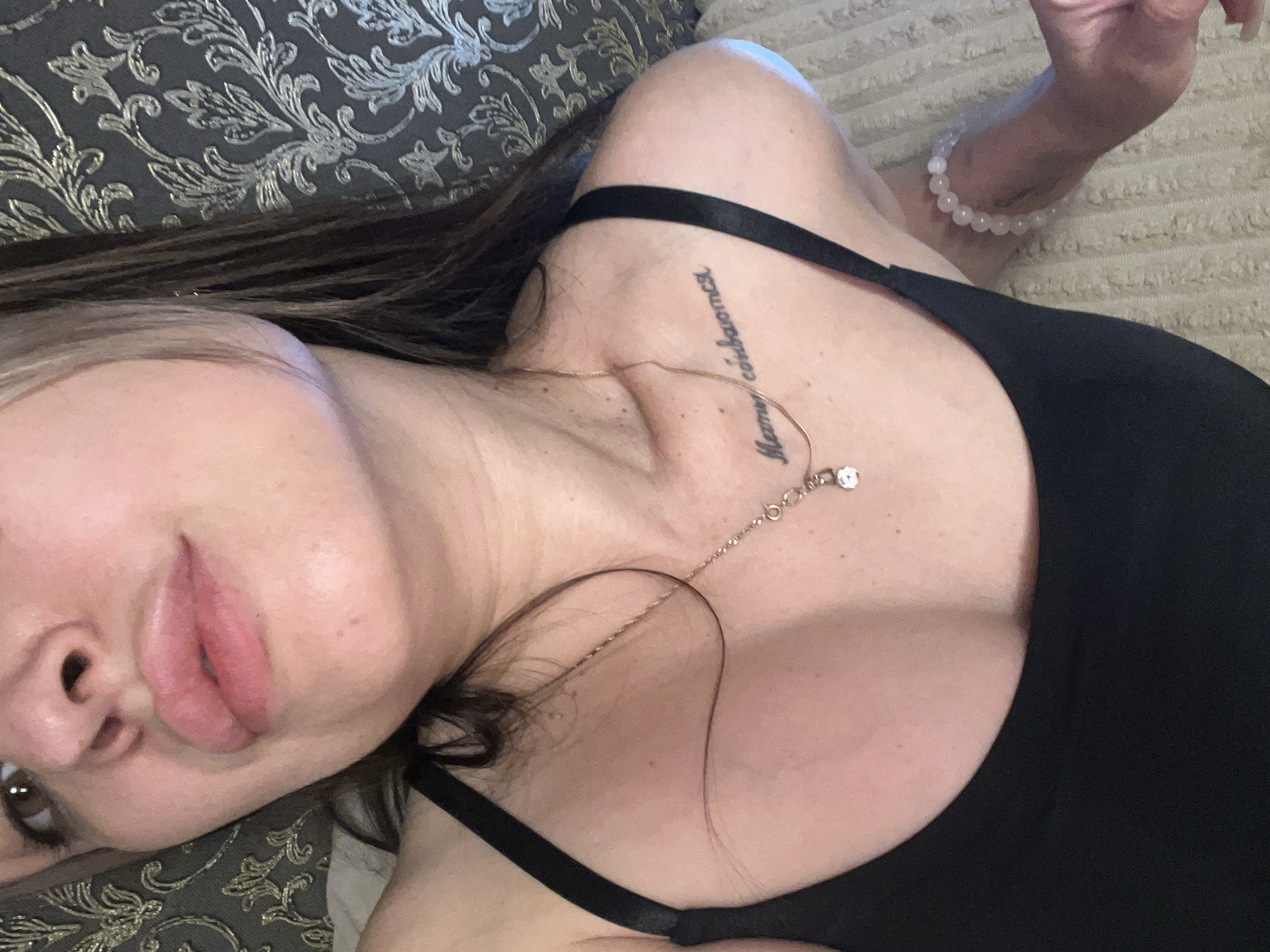 A low-angle close-up depicts a woman lying down. Her face is partially visible, featuring a prominent nose and full lips. She has brown eyes and dark hair. She is wearing a black top with thin straps. A gold necklace with a small pendant rests on her chest. A tattoo in a cursive script is visible on her collarbone. On her right wrist, she wears a beaded bracelet. She is lying on a cushion or pillow with a metallic, patterned fabric. The setting appears to be indoors. Location information is not specified and labeled as Unknown, Unknown.