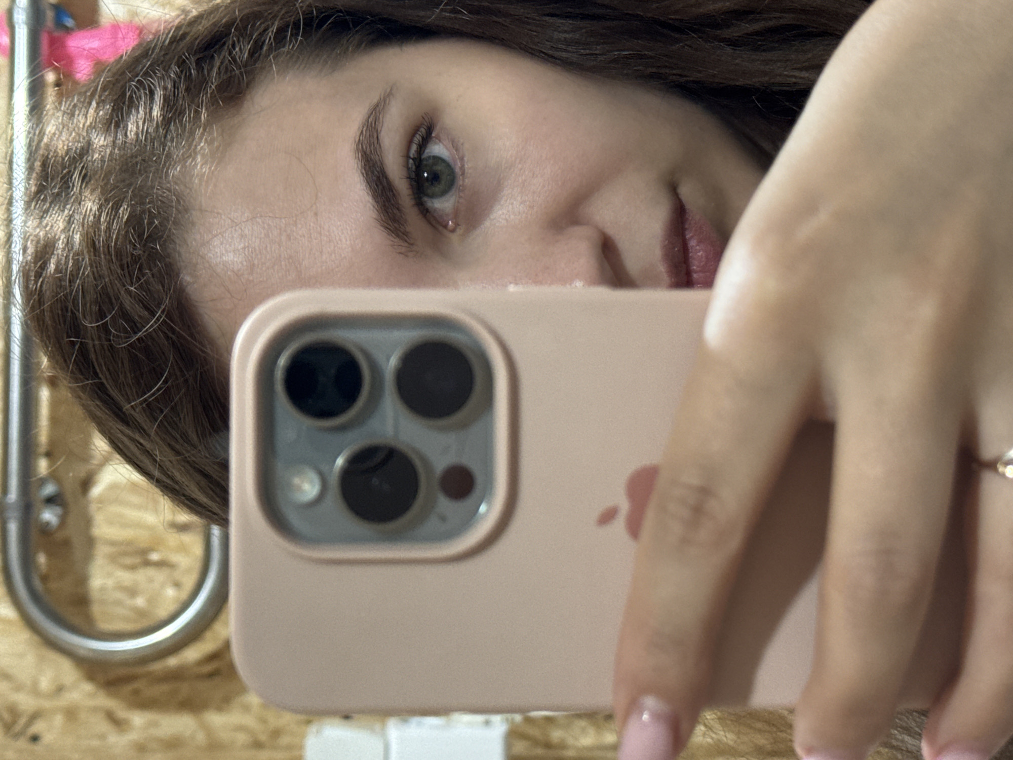 A selfie image depicts a young woman holding an iPhone. The phone, in a pink case, is held in front of her face. Her eyes are green. She has brown hair and is wearing a ring. The background shows a wooden surface and a metal bar. This image was taken somewhere near Nizhnyaya Gar', Russia, based on available data.