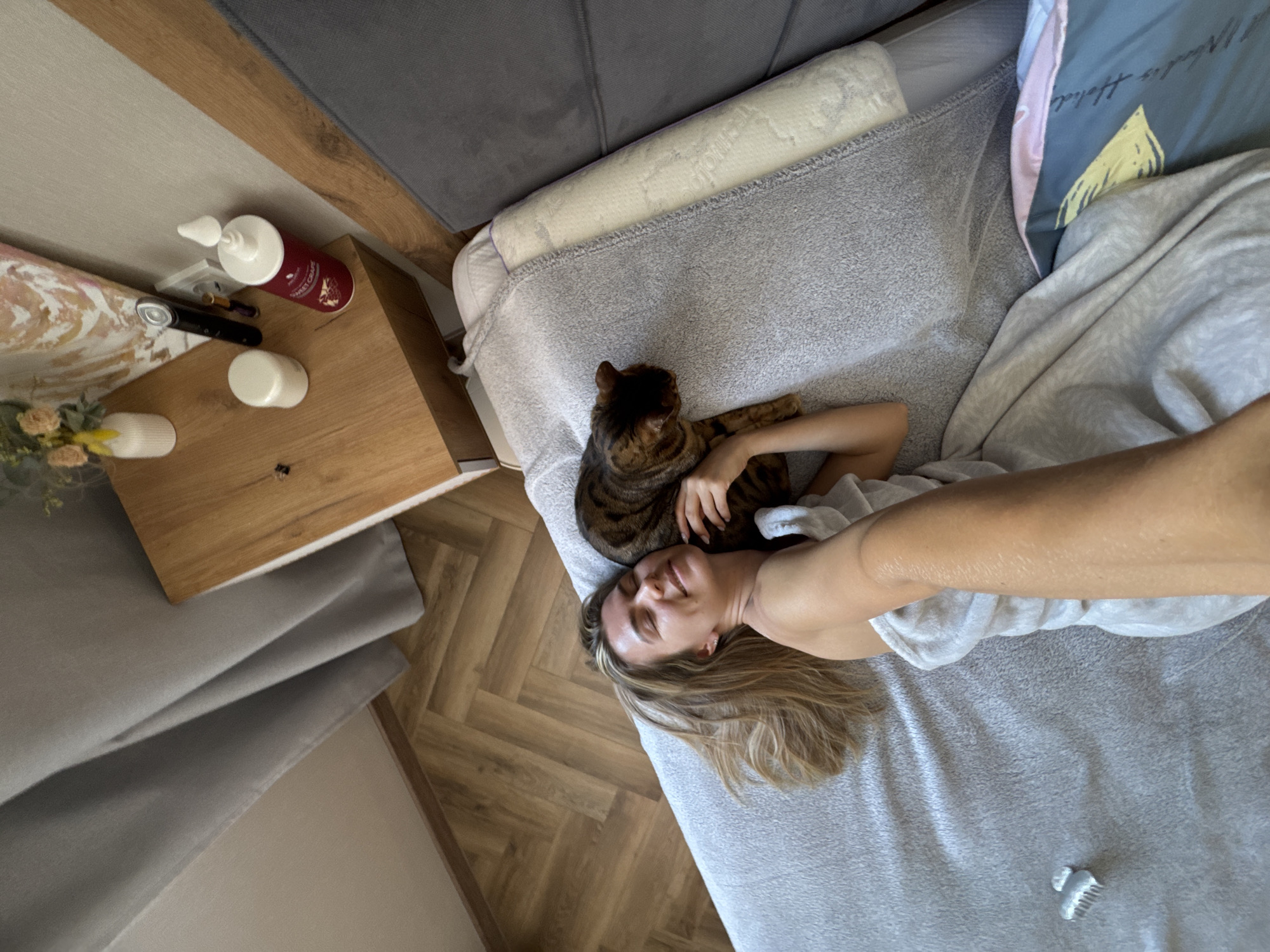A high-angle shot depicts a woman lying on a bed with a cat. The woman has light skin, blonde hair, and is wearing a white shirt. She is lying on a grey blanket. A brown tabby cat is resting on her head. To the left of the bed is a wooden nightstand with various items on it, including a white bottle, a dark red bottle, a makeup brush, and a small vase with flowers. The floor is covered with wooden planks. The location is Unknown, Unknown.