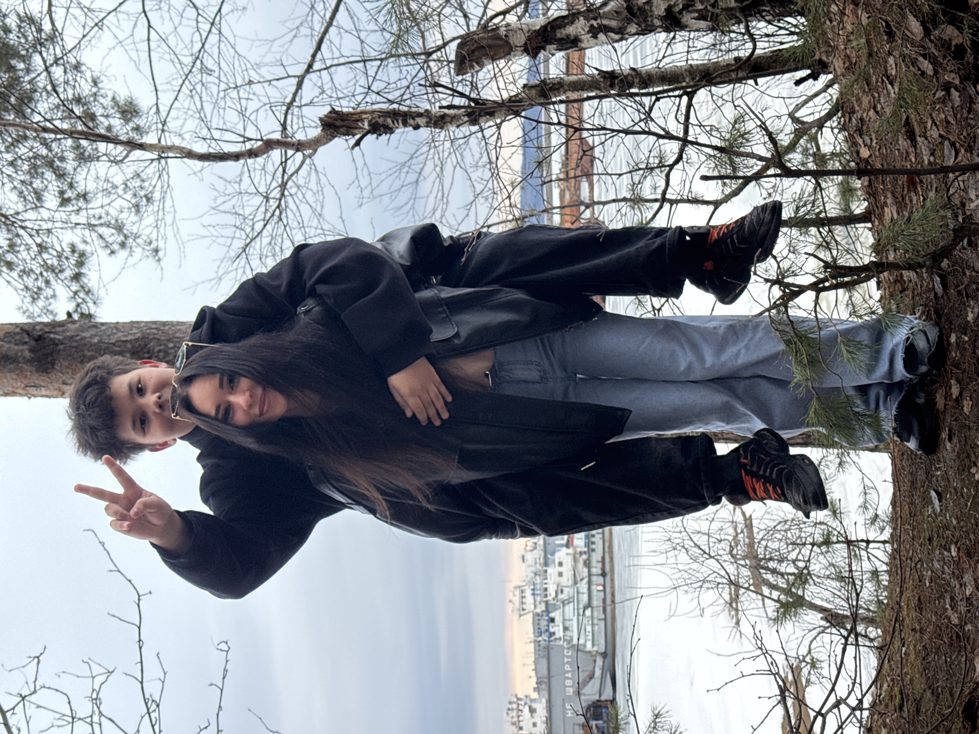 Two individuals are pictured outdoors. The individual on the left is making a peace sign. Both individuals are dressed in dark jackets, light-colored jeans, and dark shoes. The backdrop suggests a waterfront, possibly a dock or pier, with buildings and water. The surrounding area contains trees with bare branches, and some evergreen trees are visible. Based on visual clues, the location may be Chaikovsky, Russia, judging by the buildings in the background. The image shows a calm day near water.