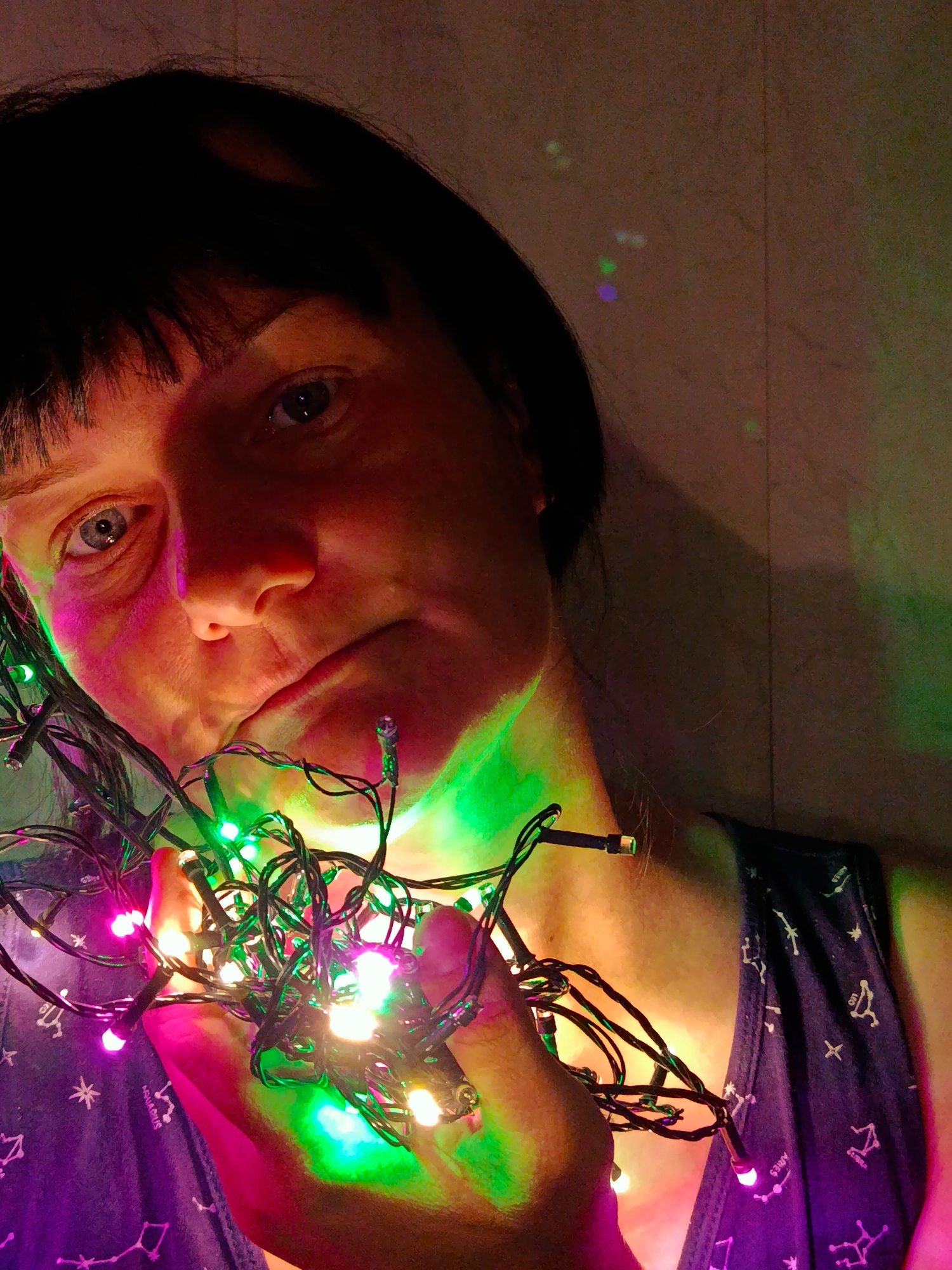 A close-up, indoor shot shows a person holding a tangled string of colorful Christmas lights. The lights cast green, pink, and white hues across the person's face and neck. The individual has short, dark hair with bangs and appears to be wearing a blue sleeveless top with constellation patterns. The background is a pale, textured wall. The location is Unknown, Unknown.