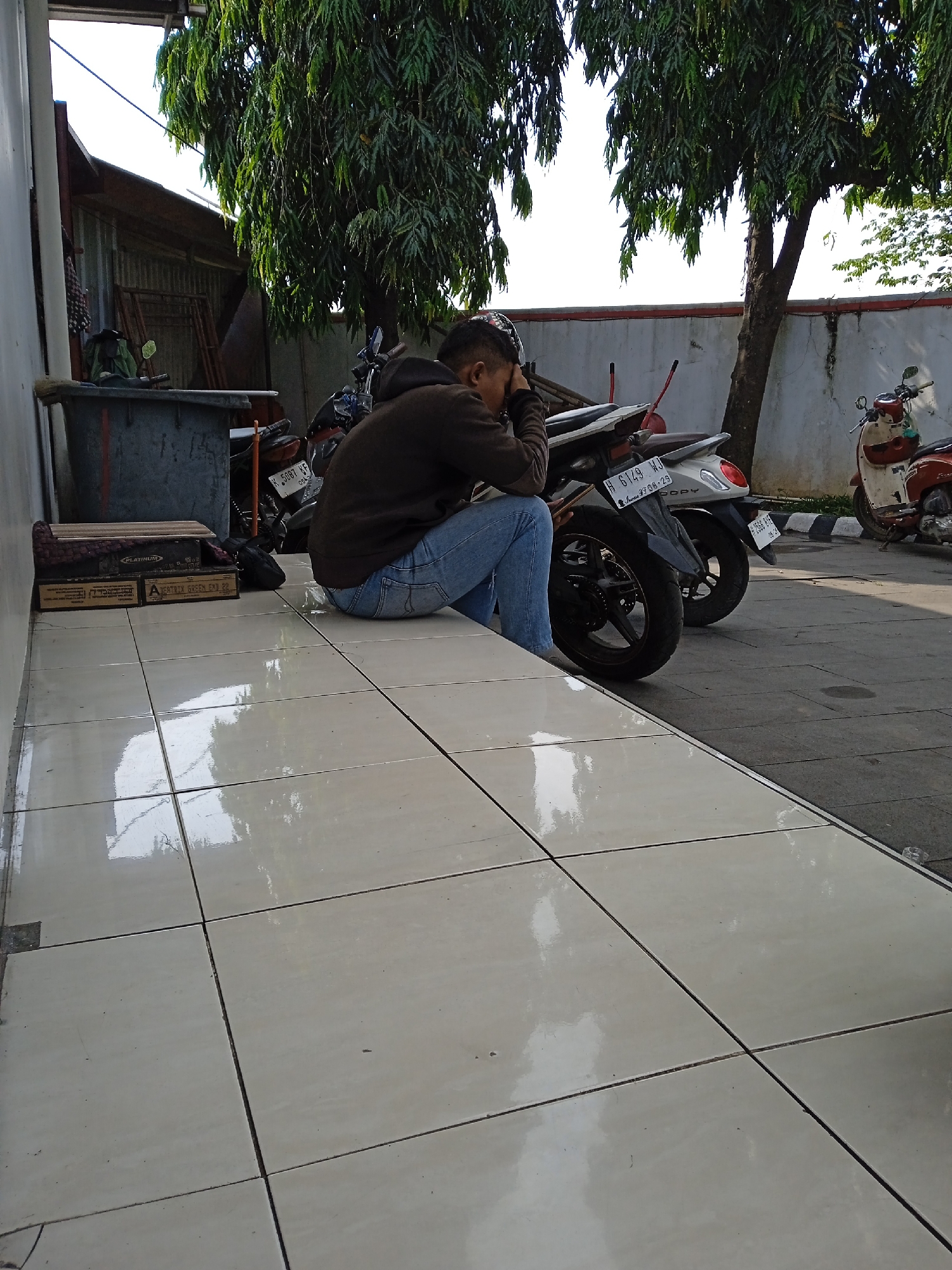 The image, taken outdoors in what appears to be Katonsari, Indonesia, shows a person seated on a tiled ledge with their head in their hand, suggesting a posture of pensiveness or weariness. The person is wearing a dark hooded top and blue jeans. To the left of the person is a grey rectangular container with miscellaneous items on top and behind it. A stack of cardboard boxes are also on the ledge, adjacent to the person.
Behind the seated individual, there are three motor scooters parked. The license plates of the scooters visible in the image have the following identification numbers: H 5087 KF and H 6149 WJ. The motor scooters are parked near a white wall with black and white striped curbing.
The surroundings are somewhat urban, with trees providing shade and an indication of daylight. The light suggests it might be mid-morning or mid-afternoon. The tiles on the ledge are light-colored and reflective.