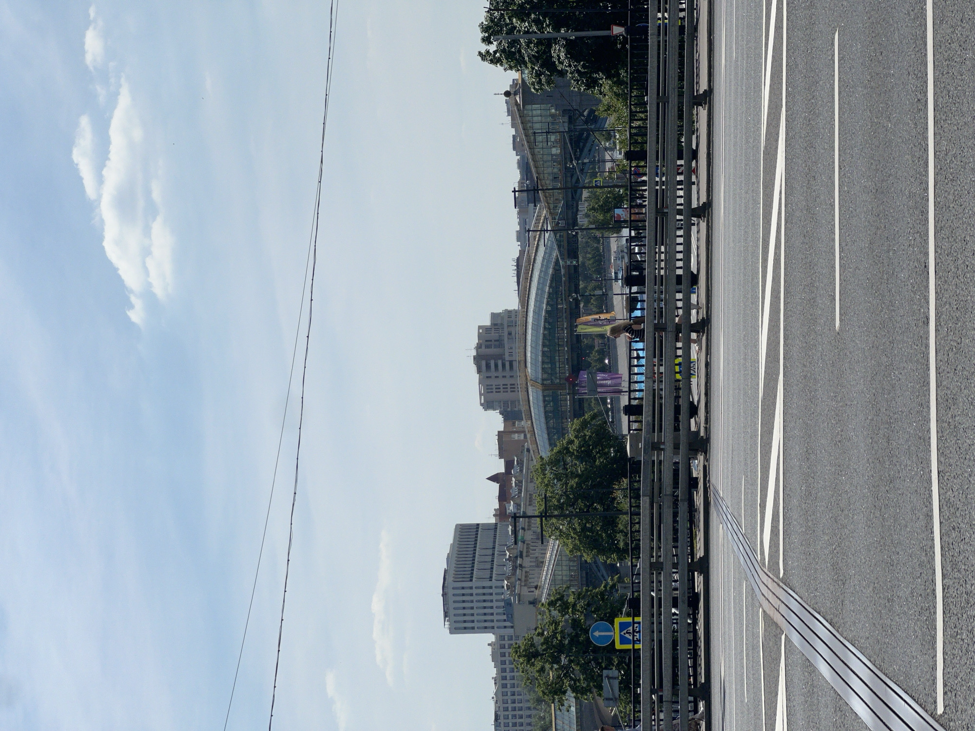 The image depicts a sunny urban scene in Moscow, Russia, observed from a road leading towards the Moscow International Business Center. In the foreground, an asphalt road surface with white lane markings and two parallel metal rails (likely tram tracks or bridge expansion joints) runs horizontally. A multi-lane road is separated from a pedestrian area by a metal traffic barrier, behind which a dark metal fence with railings is visible.
On the pedestrian path, a person with blonde hair, wearing a striped top, walks from left to right. Blue traffic signs, including an upward arrow and a pedestrian crossing symbol, are mounted on a pole to the left of the frame's center.
The midground and background showcase the Moscow skyline. Prominently featured is the Bagration Bridge, a distinctive glass-enclosed pedestrian bridge, spanning the Moscow River (though the river itself is obscured by trees and structures). To the left of the bridge, a tall, white, multi-story modern building with numerous vertical windows dominates, characteristic of the architecture in the Moscow City complex. Other buildings, some with reflective glass facades and a smaller red-roofed structure, are visible among green deciduous trees. The sky is a clear light blue with scattered white cumulus clouds, and overhead power lines traverse the upper portion of the frame.
