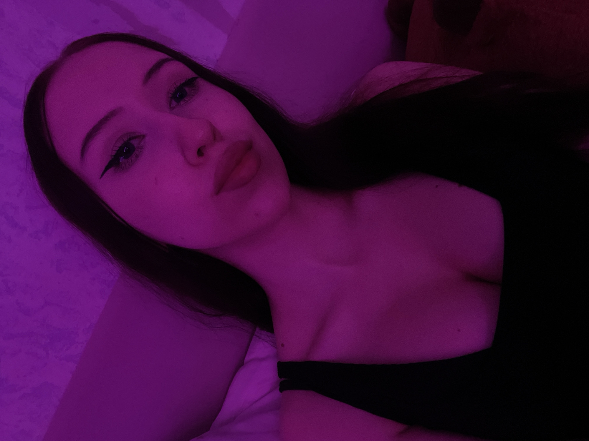 The image shows a close-up, waist-up portrait of a young woman with long, dark hair and fair skin, illuminated by purple lighting. She is wearing a black, scoop-neck top that reveals her décolletage. Her makeup includes prominent eyeliner and mascara, and her lips are full and appear to be lightly colored. The background is out of focus, but appears to be a textured wall with some indistinct shapes. The overall impression is of a selfie taken in a dimly lit room. There is no indication of Votkinsk, Russia, or any specific location in the image.