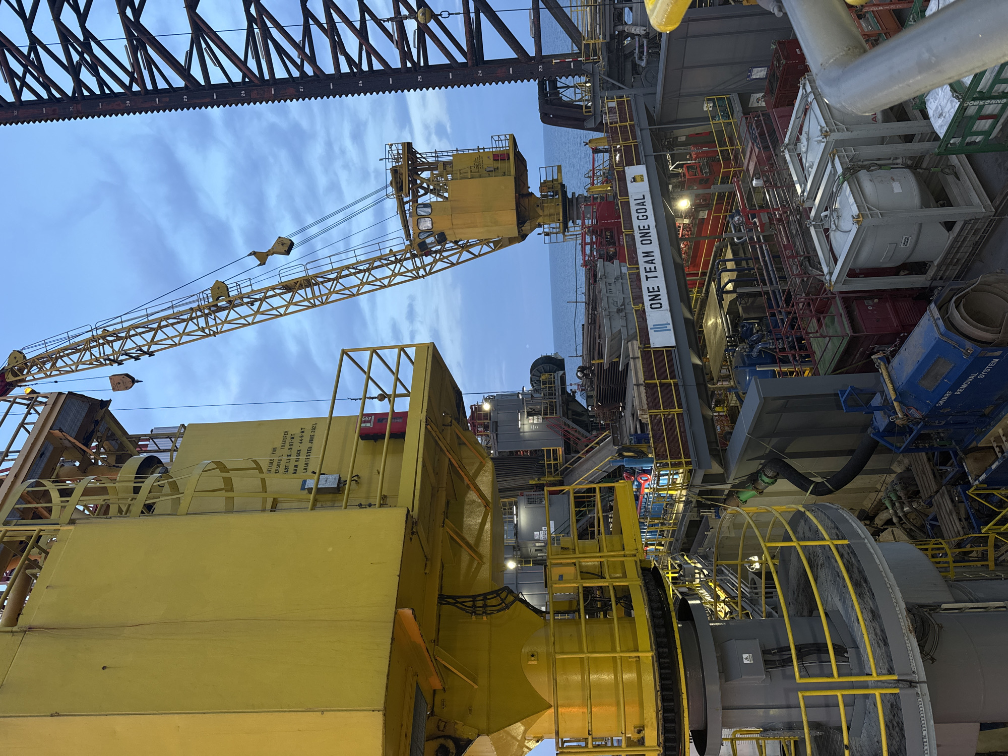 The image depicts an offshore oil rig under a cloudy sky. A large yellow crane dominates the left side of the frame, with a smaller yellow crane visible further back. Various industrial equipment, including tanks and machinery, are present on the decks of the rig. A sign on a walkway reads "ONE TEAM ONE GOAL." The sea is visible in the background. The image is taken from a high vantage point, looking down onto the rig's structure. There are no people visible in the frame, nor any indication of a specific city in Italy.