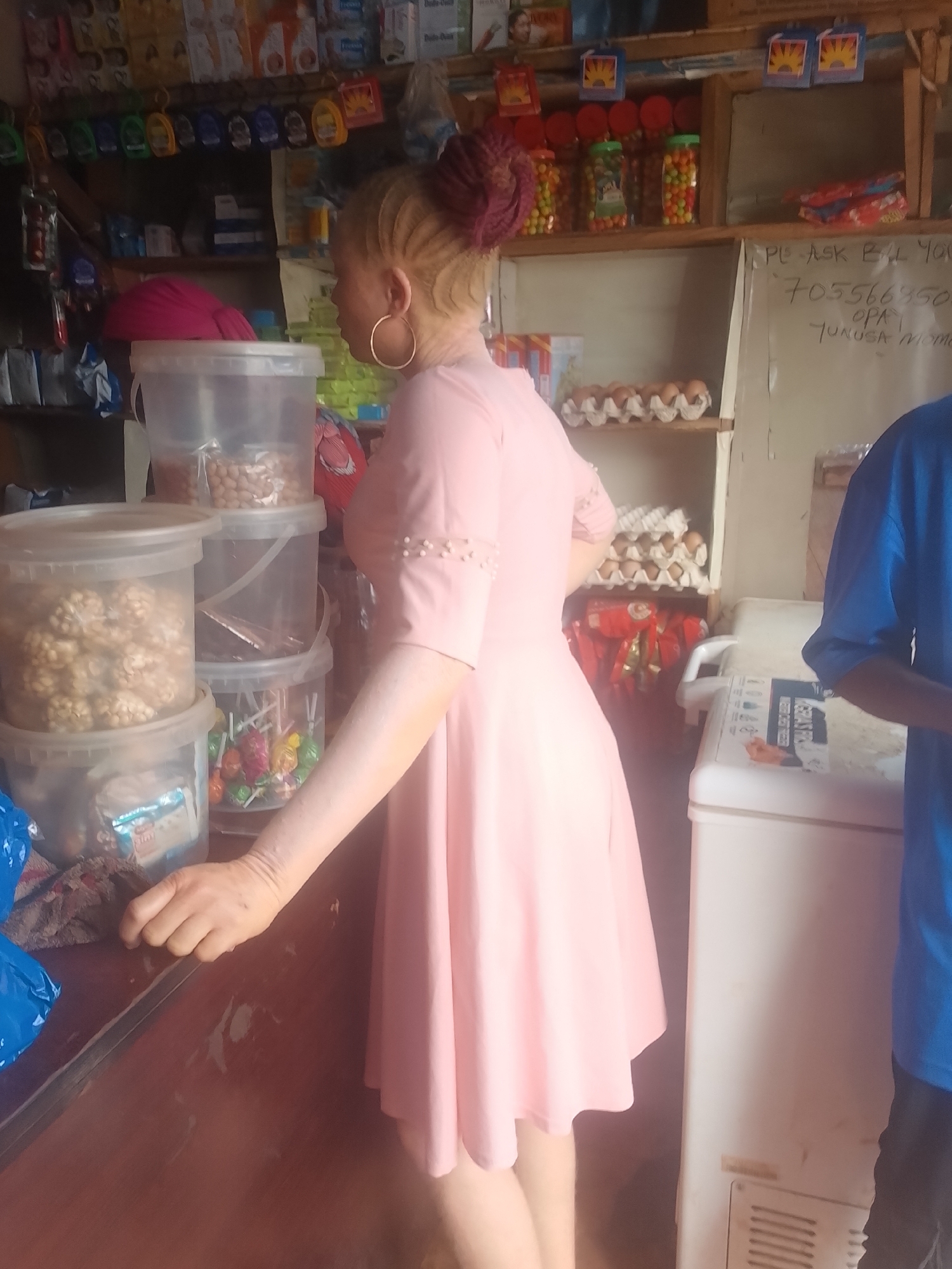 A person in a light pink dress stands in a store in Ejule, Nigeria. Shelves are stocked with various items, including candies, eggs, and assorted goods in plastic containers. A white freezer stands to the right. Another person wearing a blue shirt is partially visible. The background shows a typical local shop setting.
