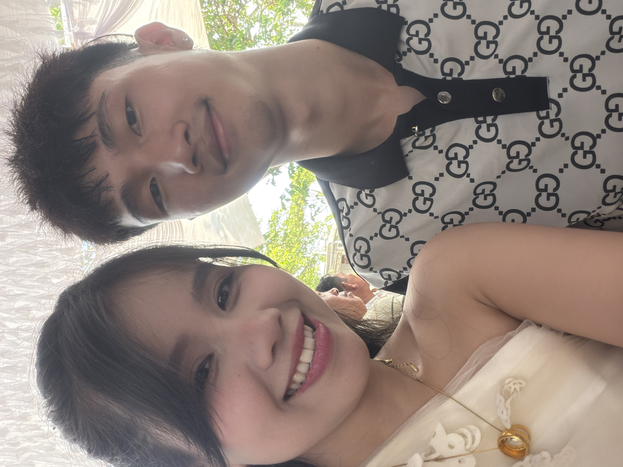 A close-up, upward-angled selfie captures a smiling man and woman, likely a couple, at an outdoor or semi-outdoor event in Bình An, Vietnam. The image is oriented vertically, with the man positioned towards the upper left and the woman in the foreground towards the lower right, both looking towards the camera with warm expressions.
The man has short dark hair and a small earring in his left ear. He is wearing a black and white polo shirt patterned with prominent "GG" logos, featuring a black collar and a placket with two visible silver buttons.
The woman has long dark hair and a wide, bright smile, revealing her teeth. She is wearing a light-colored, possibly cream or white, dress or top with subtle lacy embellishments. She is adorned with a delicate gold necklace featuring a small pendant and a gold ring on her finger.
In the blurred background, green foliage and bright natural light suggest a sunny daytime setting, likely under a white tent or canopy, parts of which are visible in the upper left. Another person's out-of-focus head is faintly visible behind the woman. The overall mood conveyed by the subjects' smiles is joyful and celebratory.