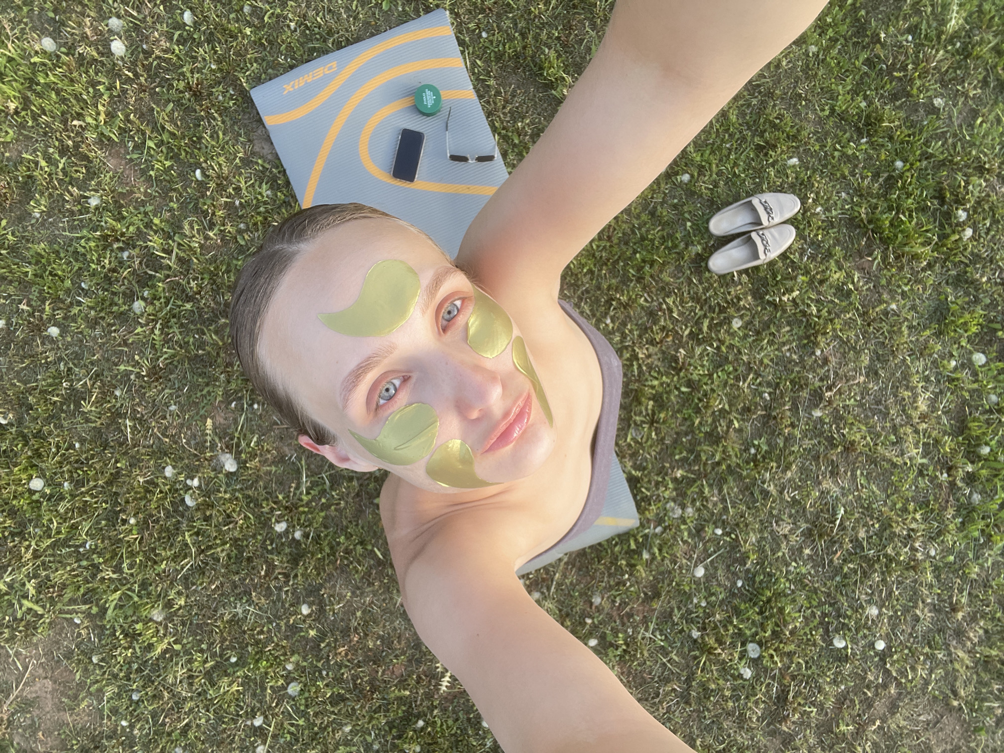 A high-angle, outdoor shot captures a woman in Подшивалово, Russia, applying gold-colored facial patches. She lies on a gray and yellow exercise mat on a grass lawn dotted with white dandelion seed heads. A mobile phone, sunglasses, and a small green jar sit atop the mat. Nearby, a pair of beige slip-on shoes rests on the grass. The woman's arms are extended upward, as if taking a selfie.