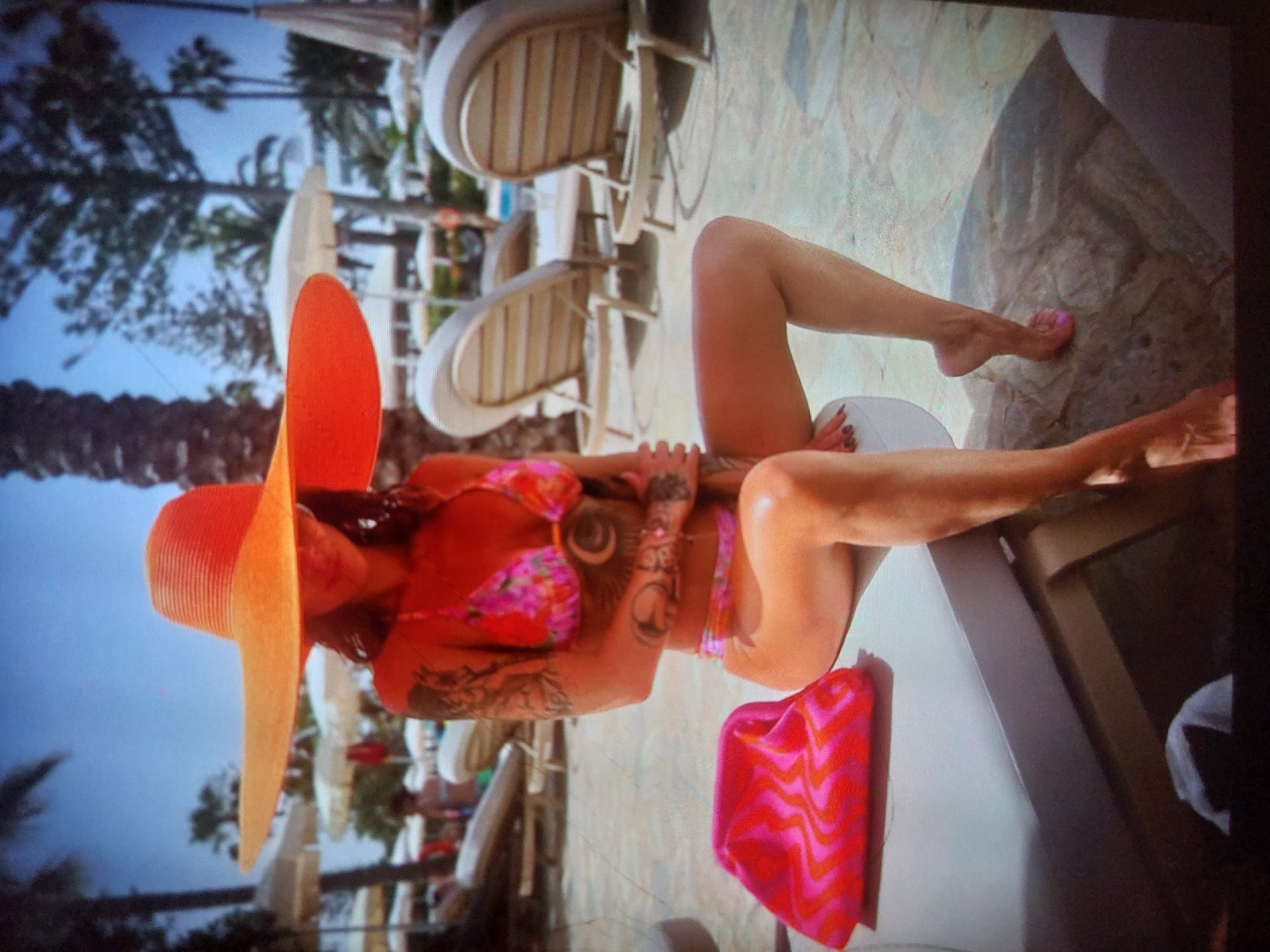 The image captures a woman relaxing outdoors in a bright, sunny, resort-like setting, identified as Tsarevo, Bulgaria. She is lying on a white or light-colored lounge chair, positioned on paved light-colored tiles or stone.
The woman is wearing a vibrant, floral-patterned bikini in shades of pink, orange, and purple, complemented by a large, wide-brimmed orange sun hat. Her long, dark hair is visible beneath the hat. She has extensive tattoos on her arms and torso, including a prominent circular design on her side. Her toenails are painted pink.
In the background, several other white lounge chairs are visible, along with numerous palm trees and what appears to be a clear blue sky, indicating a warm, tropical, or Mediterranean climate. A brightly colored pink and orange patterned towel or cloth is placed beside her on the lounge chair. The scene is well-lit by natural sunlight, suggesting it is daytime. The overall impression is one of leisure and relaxation in a vacation environment. No visible text is present in the image.