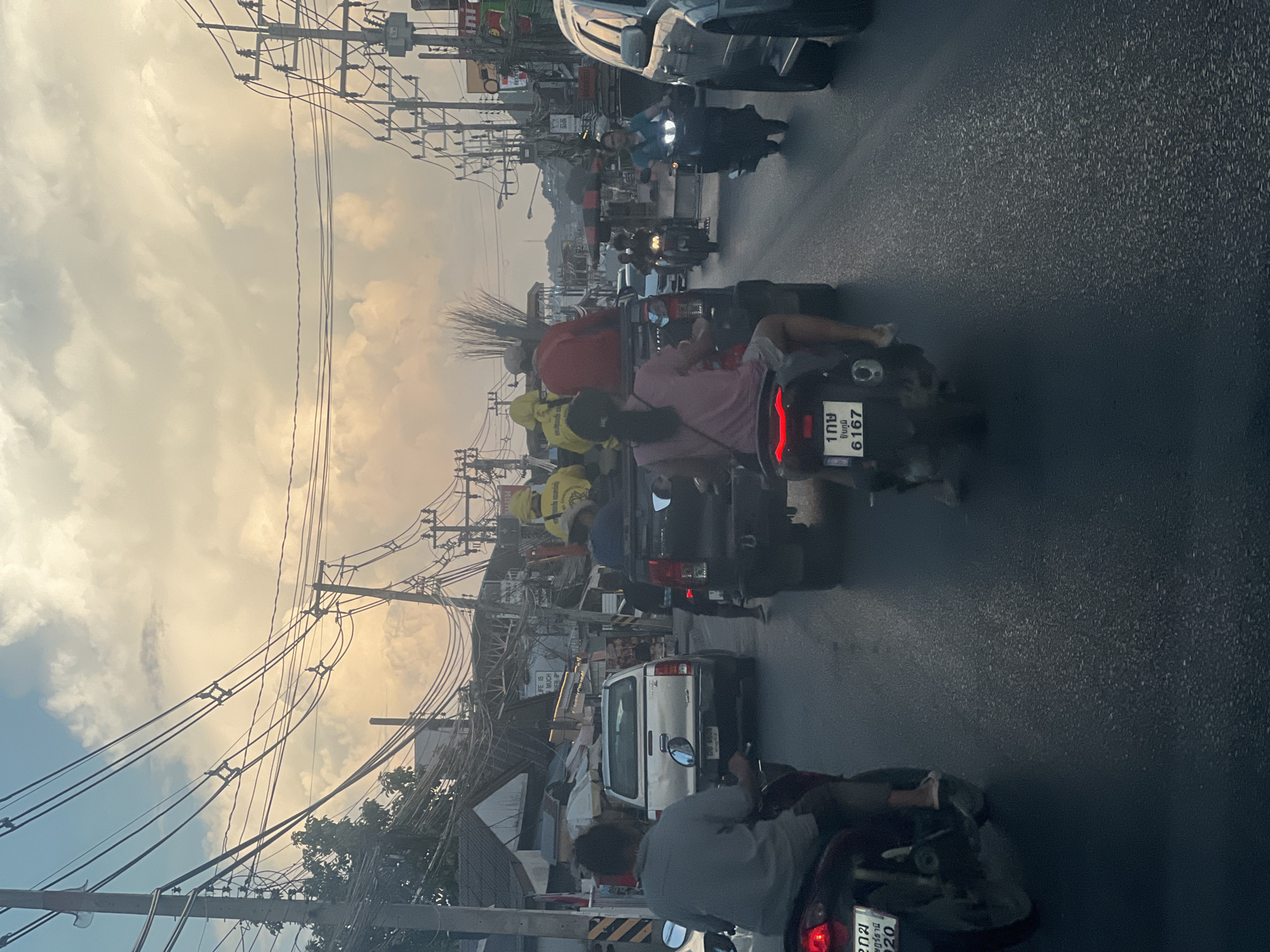 The image depicts a traffic scene on a street in Ko Samui District, Thailand, under a cloudy sky. In the foreground, two motorcycles are visible, one with a person wearing a pink shirt and shorts. Behind them, a silver pickup truck is being driven, and in the back of a black pickup truck, several people are seated, some wearing yellow vests and carrying what appear to be brooms or long sticks. Other vehicles, including a dark-colored SUV and more motorcycles, are also present in the traffic. The street is lined with utility poles and wires, and buildings are visible on the sides of the road, suggesting an urban or semi-urban environment. The overall impression is of a busy street with slow-moving traffic, possibly during the late afternoon or early evening due to the lighting conditions.