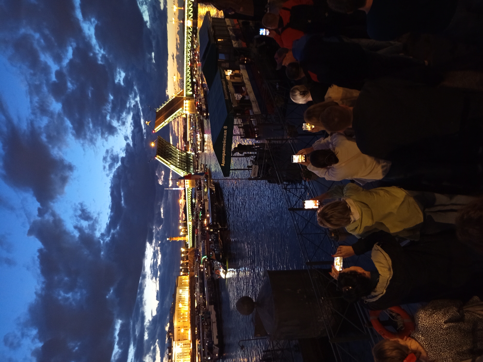 The image depicts the Palace Bridge of Saint Petersburg, Russia, during its nightly opening. The scene is set at dusk, with a deep blue sky overhead filled with dramatic, dark clouds. The bridge is raised, with its two sections forming a V-shape, illuminated by bright lights.
In the foreground, a crowd of people are gathered on a pier, facing the bridge and the Neva River. Many are holding up their smartphones, capturing the spectacle. The river is dark, reflecting the lights of the city and the bridge. Several boats, likely tour boats, are moored or moving on the river.
On the left side of the image, a building with illuminated windows is visible along the embankment, possibly part of the Hermitage Museum complex. A prominent rostral column, the Rostral Columns, also stands illuminated further down the embankment. A green awning with "Адмиралтейская 8" written on it indicates a waiting area or ticket booth for boat tours.
The overall atmosphere is one of shared anticipation and wonder, as the city's iconic landmark is revealed. The lighting suggests a transition from day to night, with artificial lights beginning to dominate the scene.