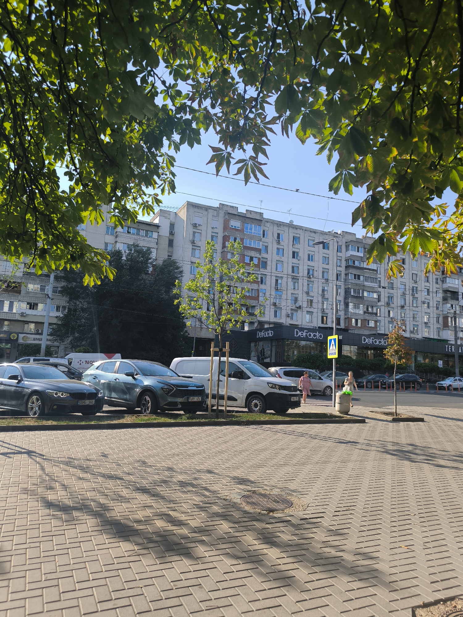 The image depicts a street scene in Chișinău, Moldova, on a sunny day. In the foreground, a paved pedestrian area with a herringbone pattern is visible, with shadows cast by tree leaves. Behind this, a row of parked cars is situated along a curb. The cars visible include a dark gray sedan, a light blue SUV, and a white van. Further back, a large, multi-story apartment building dominates the background. The building has a uniform facade with numerous windows and balconies, and air conditioning units are mounted on many of them. Several storefronts are visible at the ground level of the building, including a "DeFacto" clothing store. Two women are walking on the sidewalk in front of the building, and a pedestrian crossing signal is present. A small tree with green leaves is planted on the sidewalk. The sky is clear and blue, with some power lines visible. The overall impression is of a typical urban environment in Chișinău.