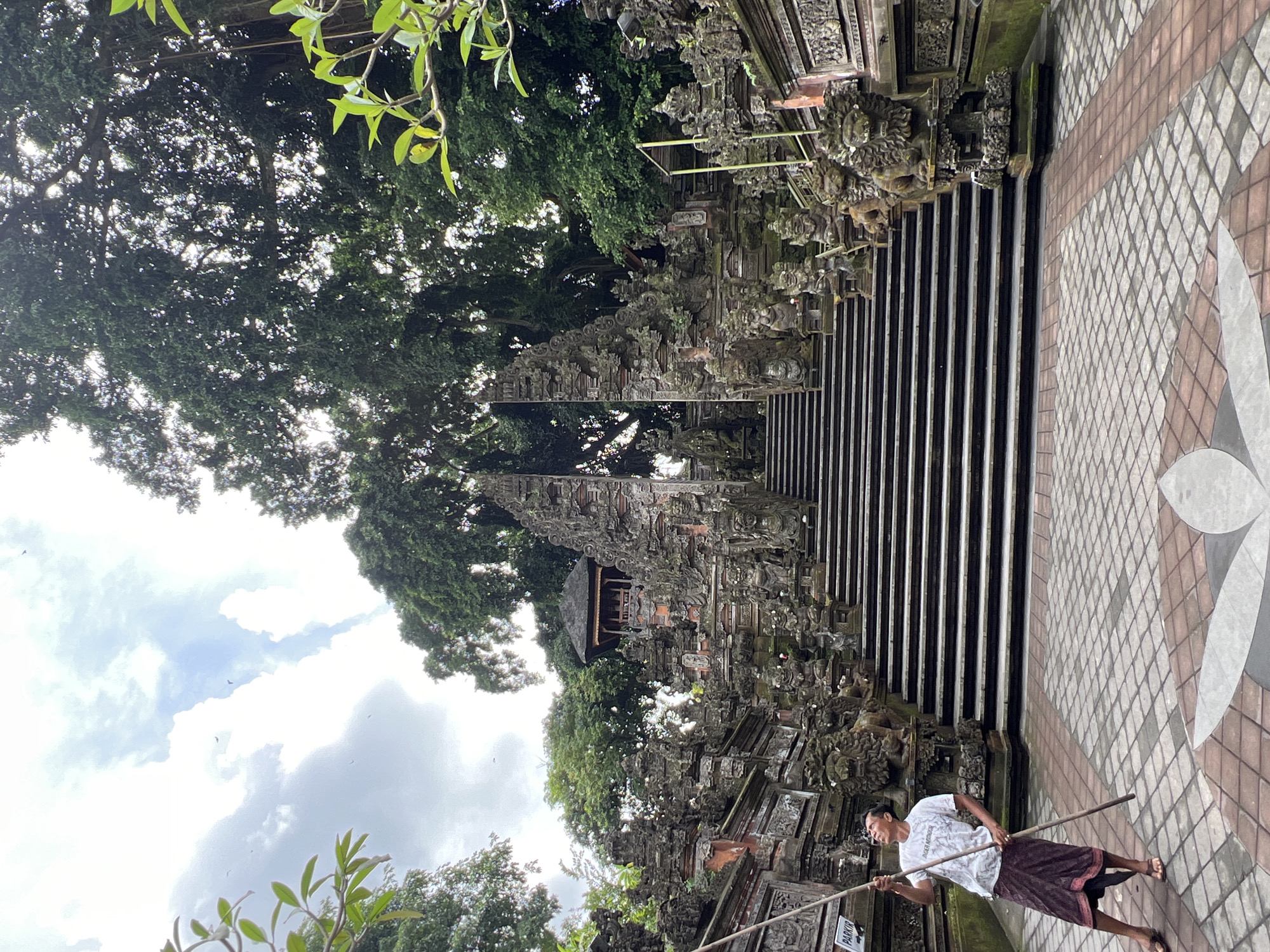 This image captures an outdoor scene at the entrance of a traditional Balinese temple or sacred site in the Ubud District, Indonesia, set amidst a vibrant tropical environment.
In the foreground, a man stands on the right side of a paved pathway, holding two long wooden sticks. He is dressed in a white t-shirt, which has faint text on it resembling "PURA MANDUG," and a dark, patterned sarong. He is barefoot and looks towards the left with a slight, content smile, appearing engaged in a task such as groundskeeping or a ritualistic activity. The paved path features a geometric pattern of brown, grey, and white tiles. In the lower left corner, a partially obscured sign appears to say "PARKIR," which is Indonesian for "parking."
The central and left sections of the image are dominated by richly detailed Balinese stone architecture. Intricately carved stone gates and multiple flights of steps ascend upwards, flanked by ornate balustrades and guardian figures carved from dark stone, some showing signs of moss and age. In the mid-ground, a small traditional pavilion with a dark roof can be seen amidst the structures.
A dense canopy of lush, towering green trees with abundant foliage frames the scene, particularly on the left and in the background, filtering the bright daytime sunlight. The sky above is partly cloudy, with bright patches where the sun breaks through. The overall impression is one of cultural richness and natural beauty.