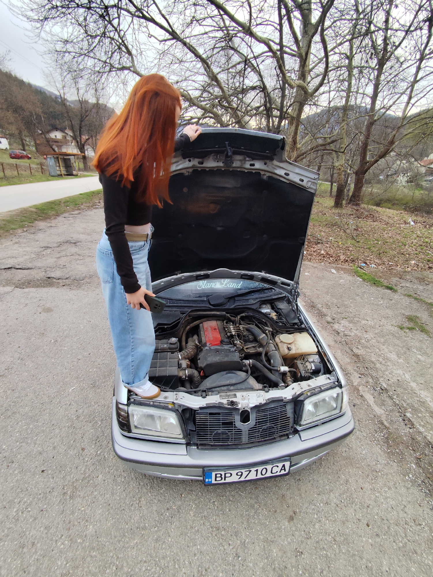A woman with long red hair stands on the hood of a silver Mercedes-Benz, which has an open hood. The woman is wearing a long-sleeved black shirt, light blue jeans, and white shoes. She is holding a black smartphone in her left hand. The license plate reads "BP 9710 CA," which is Bulgarian. The background shows a rural scene with bare trees, a road, and a building in the distance. The image appears to be taken on an overcast day. This location may be near the city of Zgorigrad, Bulgaria, based on the license plate and the landscape.