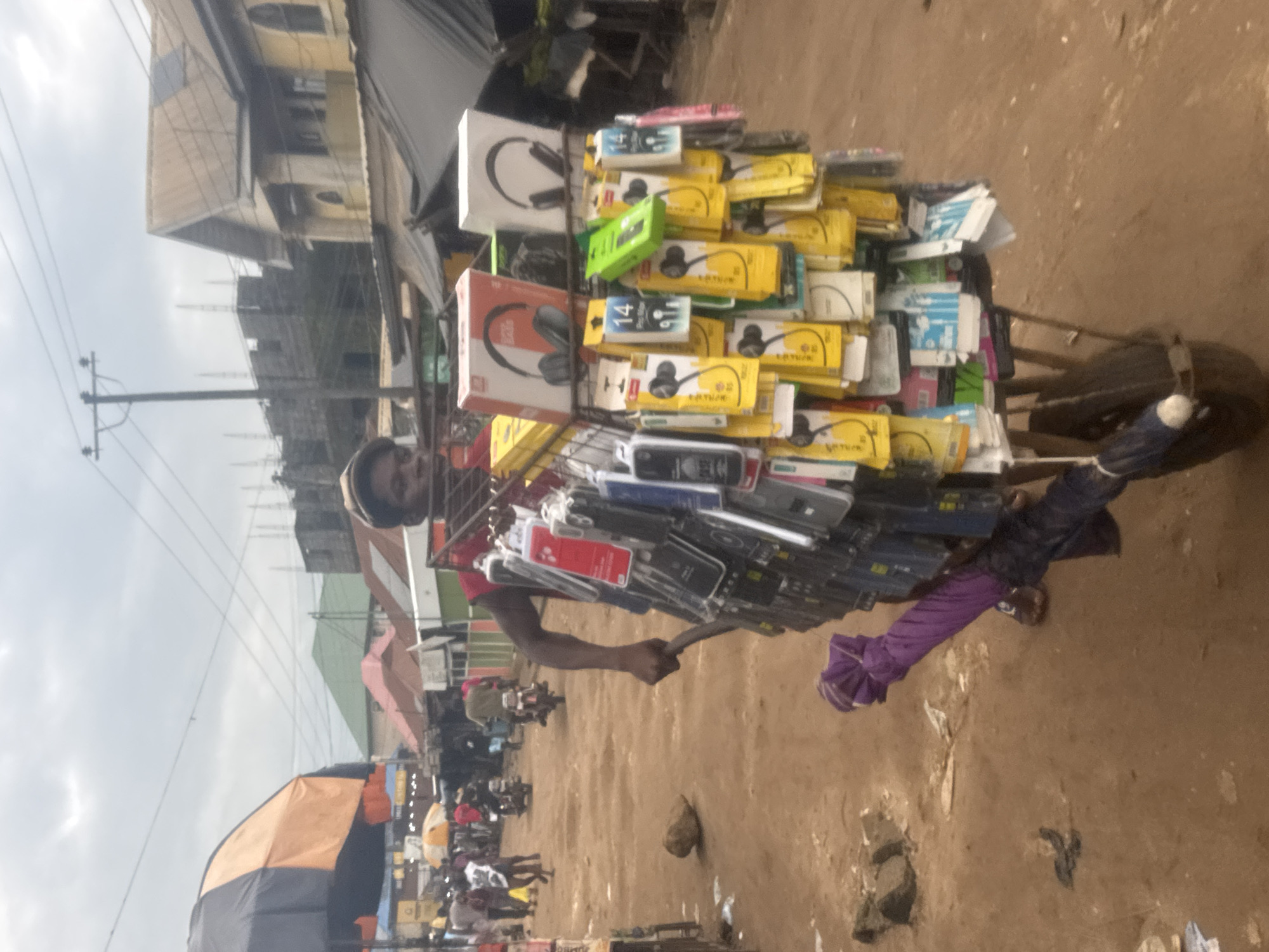 In Ihiagwa, Nigeria, a street vendor pushes a cart laden with electronics accessories through a bustling marketplace. The cart, a wheelbarrow with a metal frame, is packed with an array of items, predominantly mobile phone cases of various colors and designs, as well as numerous earphones packaged in brightly colored sleeves. Two large boxes, each displaying a picture of headphones, are prominently placed at the top of the cart. A folded purple umbrella is secured to the lower portion of the cart. The vendor, a dark-skinned man wearing a red shirt and a cap, is seen from the chest up, pushing the cart with his right hand. The marketplace is unpaved with dirt and sand, indicative of a rural or semi-urban setting. Several people are visible in the background, some walking, others on motorcycles, suggesting active commerce. Buildings with corrugated iron roofs line the sides of the street, and overhead power lines crisscross the cloudy sky. The scene captures a moment of everyday commerce in a Nigerian market.