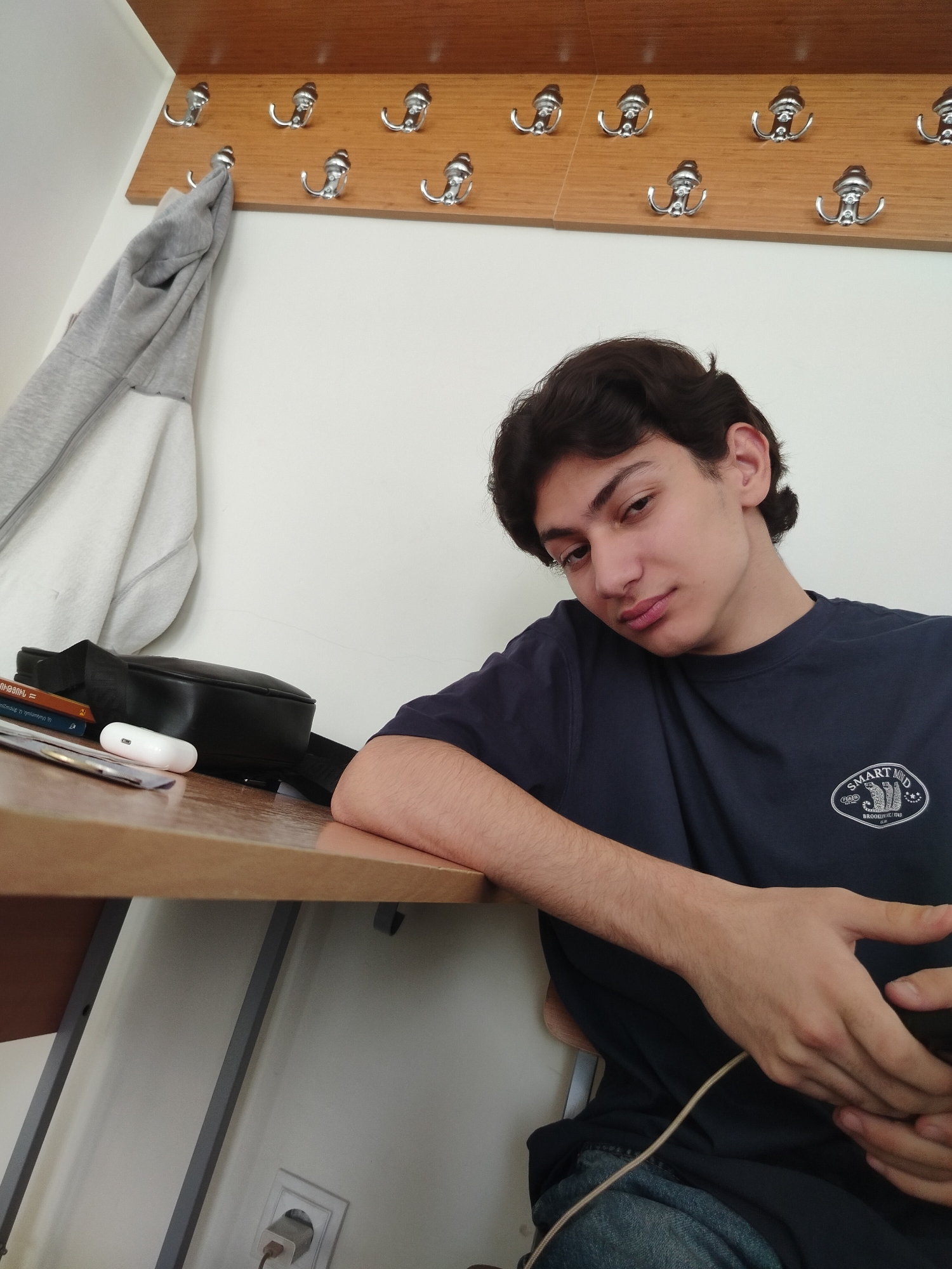 A medium indoor shot depicts a young man seated at a desk. He has dark, wavy hair and wears a dark blue t-shirt with a logo on the chest. His left arm rests on the desk, while his right hand holds a smartphone. The desk is cluttered with a black bag, a white wireless earbud case, and other indiscernible objects. Above the man, a light-colored wooden panel with several silver coat hooks is mounted on a white wall. A grey hooded sweatshirt hangs from one of the hooks. A power outlet is visible on the wall beneath the desk.
While the location is stated as Yerevan, Armenia, there is no visible information in the image to verify the location.