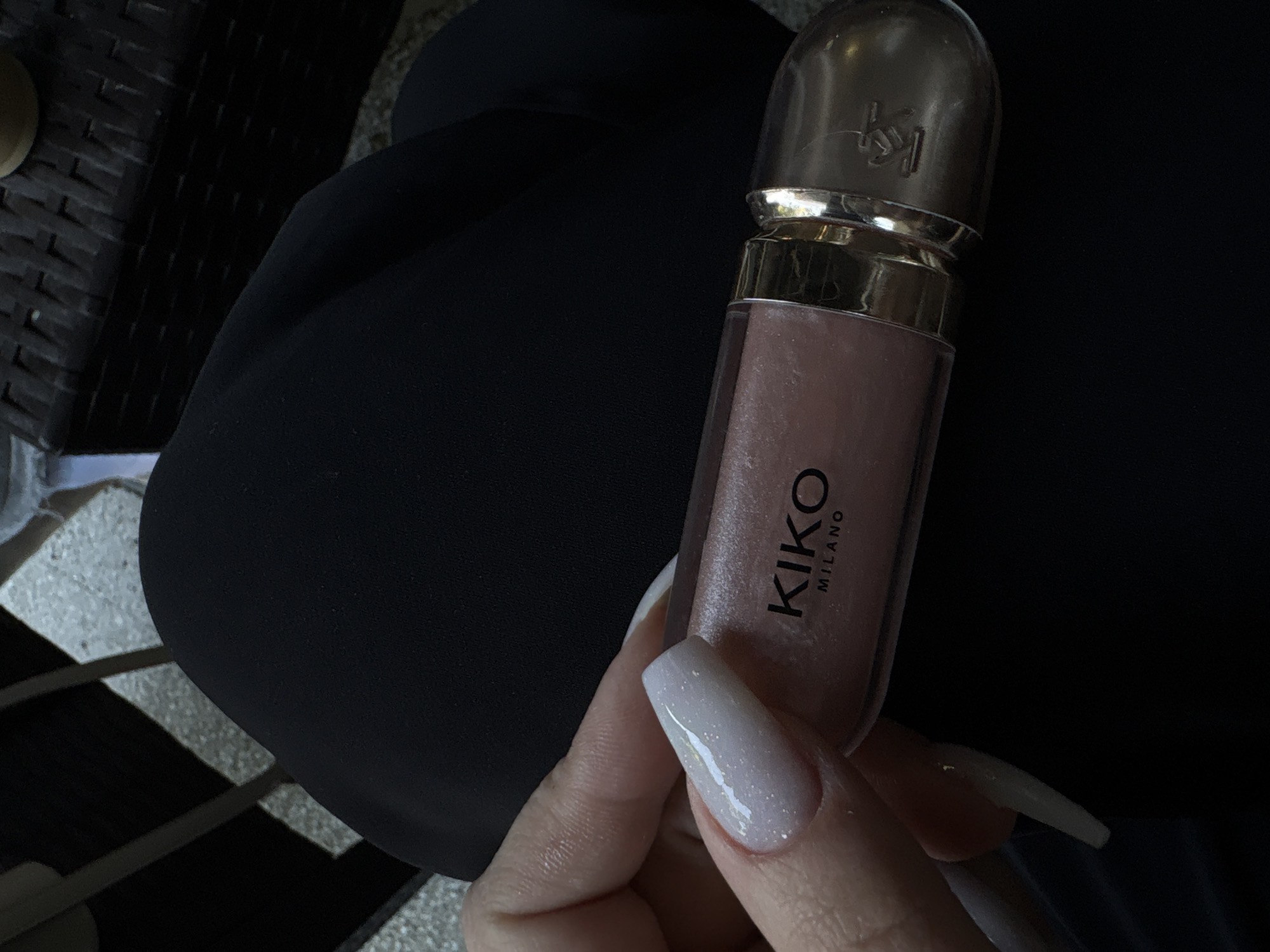 A person is holding a tube of KIKO Milano lip gloss. The lip gloss has a gold-colored top and a light pink color. The person's nails are light pink with glitter. The background is a black fabric material and a woven brown surface. The location is unspecified, but the brand of the lip gloss indicates the image may have originated in a city where the brand is sold. Gotse Delchev, Bulgaria is given in the instructions, but nothing in the image confirms this location.