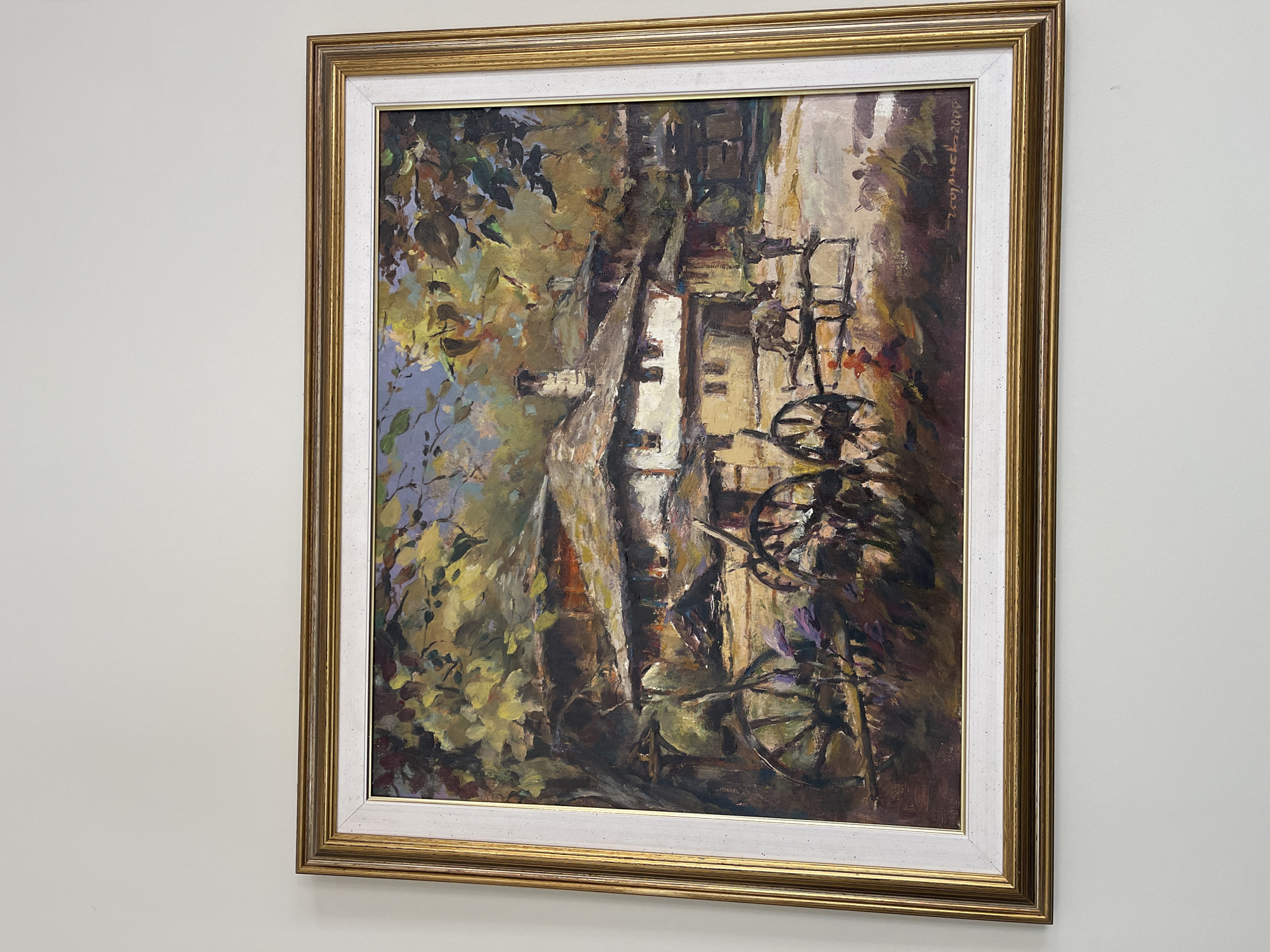 A framed painting hangs on a pale wall. The painting depicts an outdoor scene, seemingly a building façade and tree, rendered in an impressionistic style. Visible elements include architectural structures, foliage, and what appears to be a bicycle. The location, Paiporta, Spain, is not readily evident from the painting itself without external contextual clues. The painting's style is characterized by loose brushstrokes and a muted color palette dominated by browns, greens, and whites. The gold and white frame is ornate and of classical design.