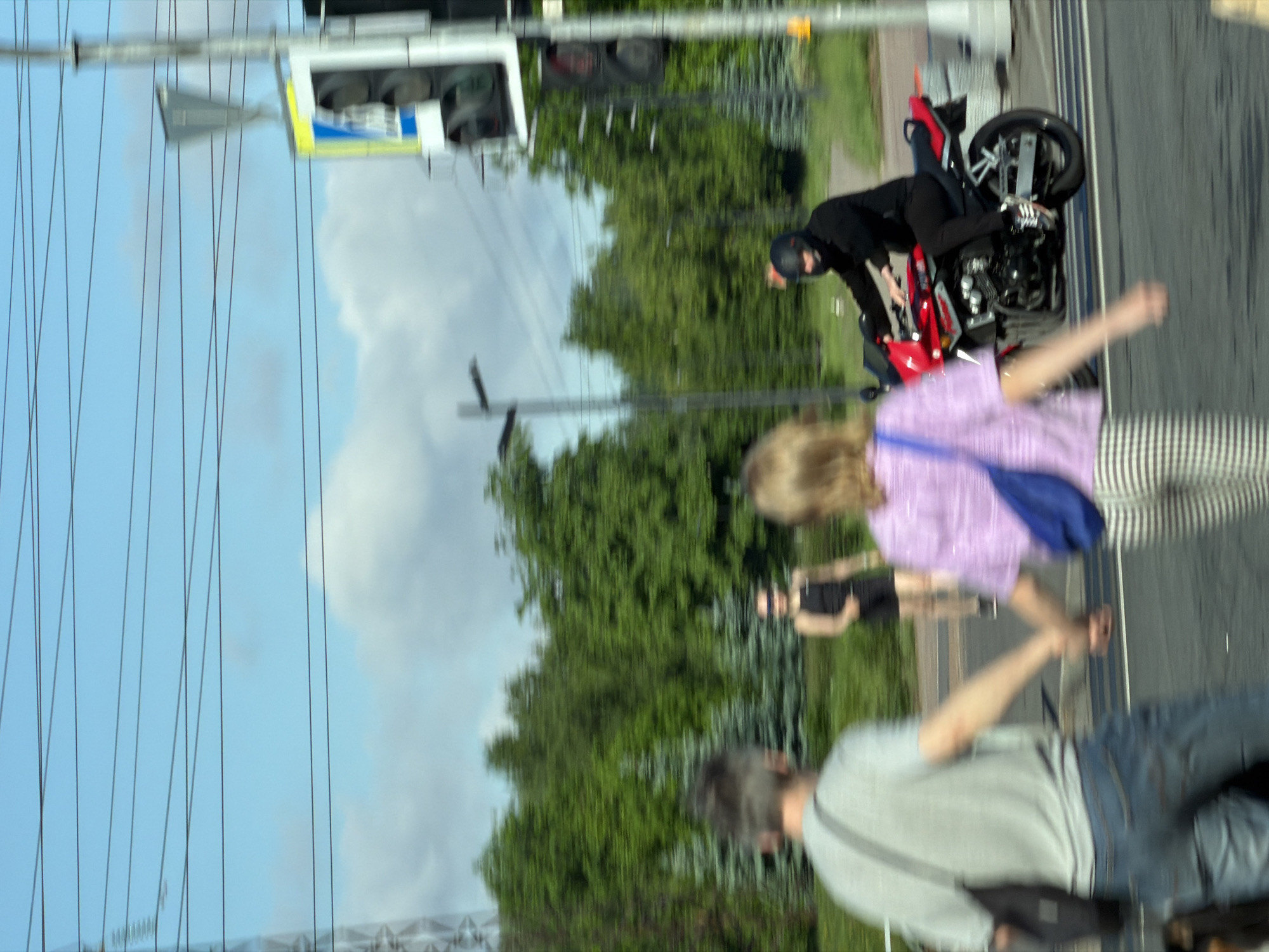 This forensic description details a daytime outdoor scene captured in the city of Tokkari-Lend, Russia, exhibiting significant motion blur throughout.
**Environmental Details:** The sky is a light blue with scattered white clouds. Multiple dark power lines horizontally traverse the upper frame. A tall metal structure, possibly a power transmission tower, is partially visible on the far left. The background consists of a dense green treeline, including both deciduous and coniferous trees, suggesting a park or forested area. An asphalt road surface with faint white markings occupies the lower portion of the frame, bordered on the right by a concrete sidewalk or median with a yellow element. A light pole stands on the left side of the road, and a tall pole with multiple traffic lights and a blue and yellow sign (likely a pedestrian crossing indicator) is visible on the far right.
**Subjects and Actions:**
1. **Foreground Couple:** Two individuals, a male and a female, are walking from right to left across the lower-left portion of the frame, holding hands. The male, on the left, wears a light gray t-shirt, blue jeans, and carries a dark shoulder bag. His head and upper body are visible, with short dark hair. The female, on the right, wears a light purple short-sleeved top, striped black and white pants, and carries a blue shoulder bag. Her face is obscured. Both are highly blurred, indicating movement relative to the camera.
2. **Motorcyclist:** A person on a red and black motorcycle is depicted in the midground, moving from right to left. The rider wears a black helmet, a black long-sleeved top, and black pants. The motorcycle features a visible engine and two wheels, with the rear wheel having a silver rim. The rider's right foot is on a footpeg. This subject is also significantly blurred, indicating motion.
3. **Third Pedestrian:** In the midground, further behind the foreground couple and to the left of the motorcyclist, a third individual is visible. This person appears to be walking from right to left in a grassy area. They wear a dark, sleeveless top and dark shorts. A dark band covers their eyes, resembling a blindfold or specific eyewear. This individual is also motion-blurred.