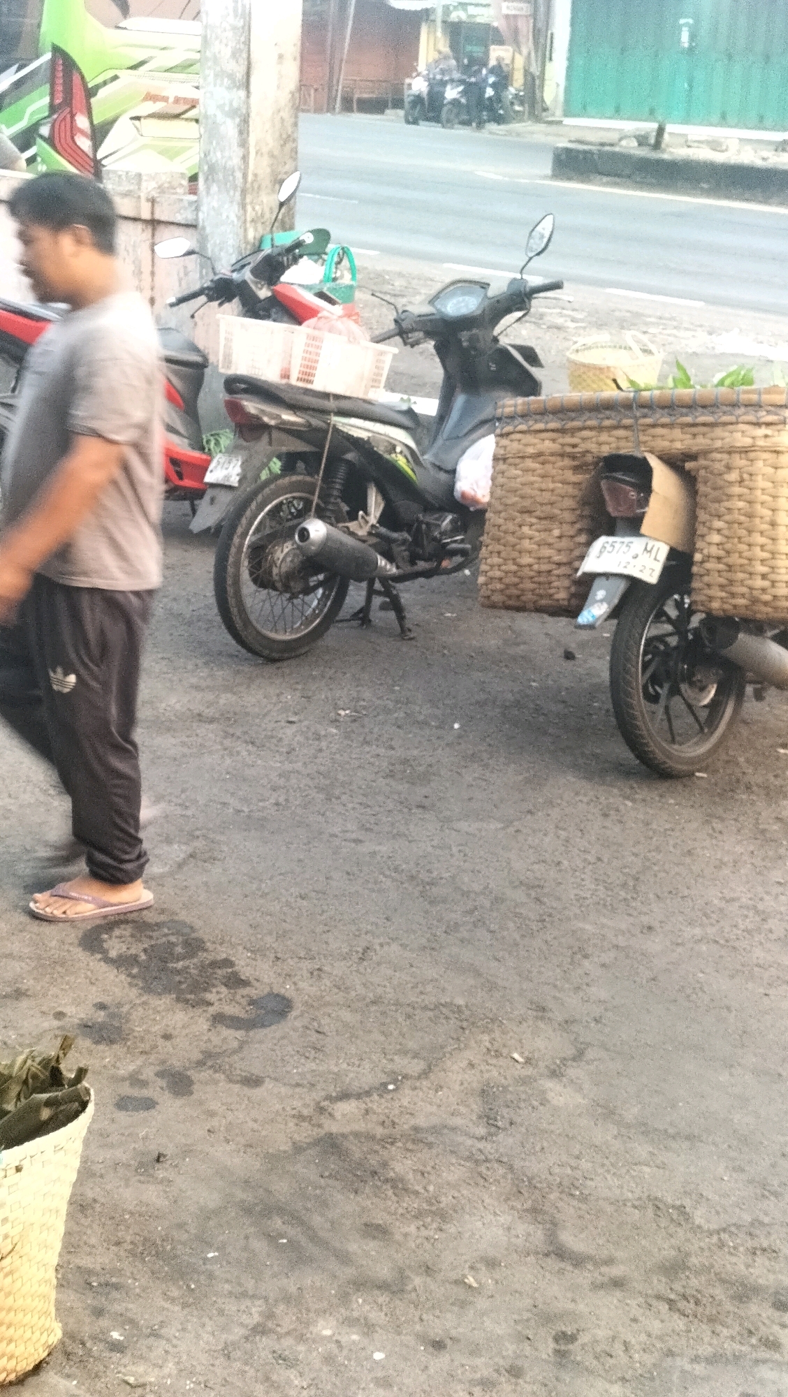 The image captures a street scene in Subah, Indonesia, likely in the morning based on the lighting. In the foreground, a person wearing a grey t-shirt and dark track pants with white stripes is walking from left to right, partially out of focus. They are wearing light-colored flip-flops. To the left, a woven basket is visible.
In the middle ground, two motorcycles are parked. The motorcycle on the left has a white basket attached to its rear. The motorcycle on the right is equipped with a large, boxy wicker basket that covers its rear wheel and part of the side. This basket has a cardboard insert. A license plate on the right motorcycle reads "B575 ML" with a smaller date below. The motorcycles are parked on an asphalt surface, which appears somewhat worn and has some dark stains.
In the background, other motorcycles are visible parked on the side of a road. Buildings and a green facade are also present. The overall environment suggests a commercial or roadside area.