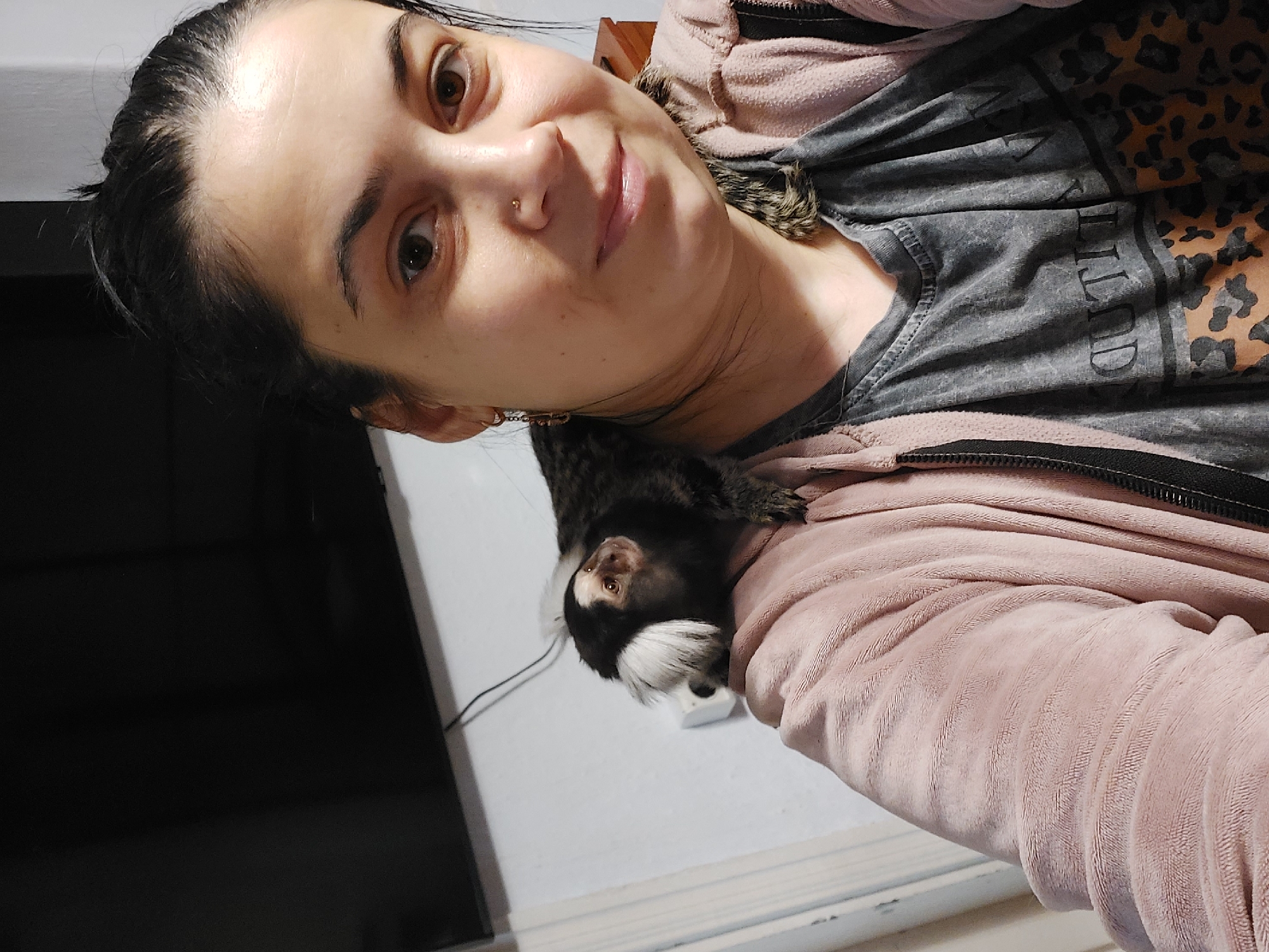 A medium close-up, eye-level indoor photograph taken in the city of Unknown, Unknown, shows a woman and a small monkey. The woman, positioned slightly right of center, has dark hair pulled back, brown eyes, and is looking directly at the camera with a faint smile. She has a small gold stud piercing in her right nostril and a small, dark earring in her left ear. She is wearing a grey t-shirt with partially visible dark lettering and a leopard print pattern on the lower portion, beneath an open, pink, soft-textured zip-up hoodie. A Common Marmoset, identifiable by its white ear tufts and facial markings, is perched on her right shoulder, facing left and looking slightly upwards. The monkey's body is predominantly dark brown and black. The background to the left features a large, dark, rectangular object, likely a television screen, with a visible power cord extending downwards. A white wall with a white electrical outlet or switch is visible behind the monkey. To the far right, a small portion of a wooden furniture piece is discernible.