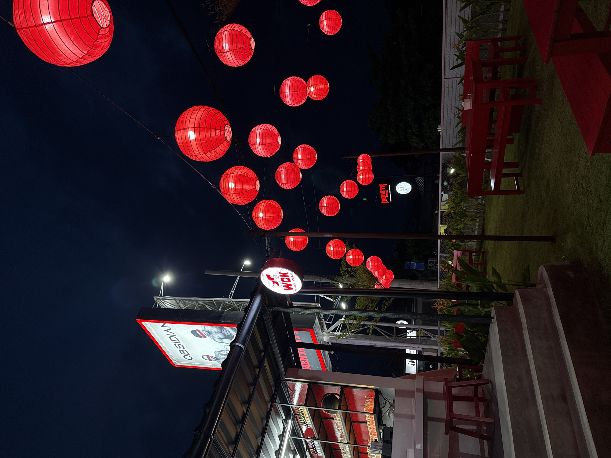 The image shows an outdoor restaurant scene at night in Munggu, Indonesia. Strings of red Chinese lanterns are strung above the premises. The restaurant has red tables and benches on a grassy lawn. The restaurant signage indicates "WOK this way" and "OBSIDIA". Stairs lead up to the restaurant entrance.