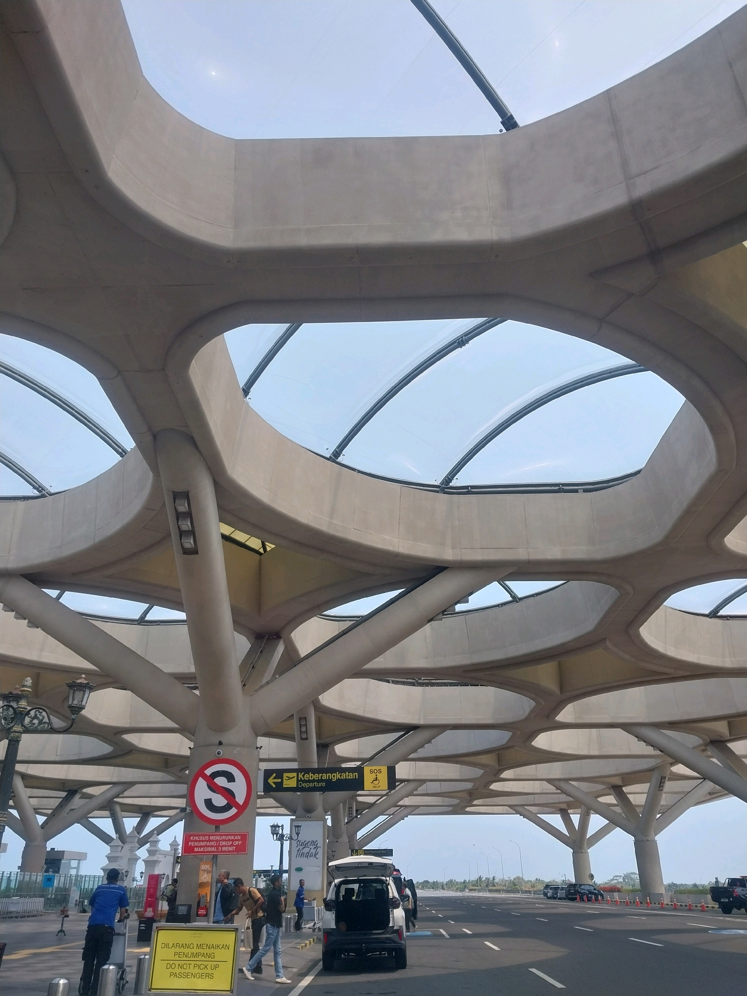 In Palihan, Indonesia, an exterior view of the Yogyakarta International Airport (YIA) showcases its distinctive canopy structure. The canopy, constructed from concrete, features an organic, honeycomb-like design with large, oval openings that allow natural light to filter through, revealing a clear sky above. Supporting this structure are numerous thick, angled concrete columns that branch out upwards, creating a visually striking architectural element.
On the ground level, a road runs parallel to the airport terminal. Several vehicles are visible, including a white SUV with its rear door open and a dark pickup truck, suggesting active passenger drop-off and pick-up activities. A yellow sign with black text that reads "DILARANG MENAIKAN PENUMPANG DO NOT PICK UP PASSENGERS" is positioned prominently near the road. This sign, along with a circular red and white prohibition sign with a stylized "S", indicates restricted areas.
A few individuals are present. One person in a blue uniform is seen pushing a luggage trolley. Another group of three men are standing and interacting. A person in a blue shirt and dark pants stands near the luggage trolley, while two other men, one in a light brown shirt and the other in a dark shirt and blue jeans, are engaged in conversation. In the background, more vehicles and people can be observed, hinting at the typical operational bustle of an airport. Traffic cones are also visible along the side of the road, potentially delineating traffic flow or restricted zones. The overall scene captures the modern architectural design of the airport and the ongoing passenger services.