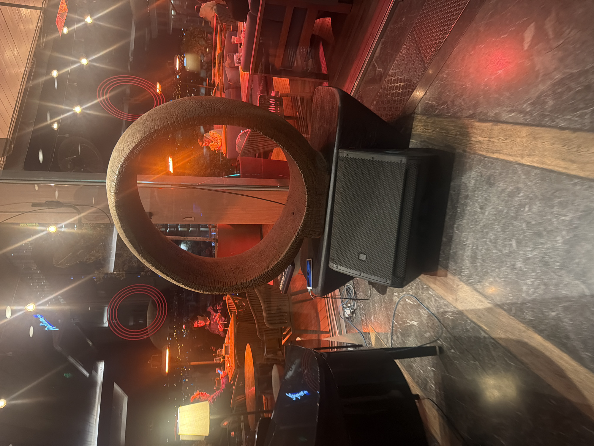 This image captures an indoor scene, likely a bar, restaurant, or lounge, viewed through a reflective glass window at night. The perspective includes elements from both inside and outside/reflected on the glass. The image is vertically oriented (rotated 90 degrees clockwise from typical landscape).
In the foreground, dominating the left side, is a large, circular, textured object made of a dark brown, possibly woven material. To the right of this, a black speaker with a mesh grille is positioned, showing the "JBL" logo on its side. A white smartphone is plugged in and resting on a surface near the speaker. The floor in the immediate foreground appears to be a dark, patterned tile.
Through and reflected on the glass, the interior of the venue is visible, bathed in warm, ambient lighting with predominant orange and red hues. Numerous light sources, some creating starburst reflections, are scattered across the image. Two distinct sets of red concentric circles are reflected on the glass, possibly from a decorative light fixture or neon sign. Faint blue neon-like text, possibly reading "Restaurant" or a similar word, is also reflected on the left.
The background shows various seating arrangements, including tables and comfortable lounge-style chairs and sofas with cushions. Several people are present, socializing or seated. One person in a dark outfit is partially visible behind what appears to be a black piano or keyboard on the lower right, suggesting a live music performance or DJ setup. Another person in a light-colored top is seated on a sofa in the upper right background. The general activity seems to be social gathering or enjoying entertainment. The time of day is clearly nighttime due to the darkness outside and the illuminated interior. The overall atmosphere appears cozy and engaging.