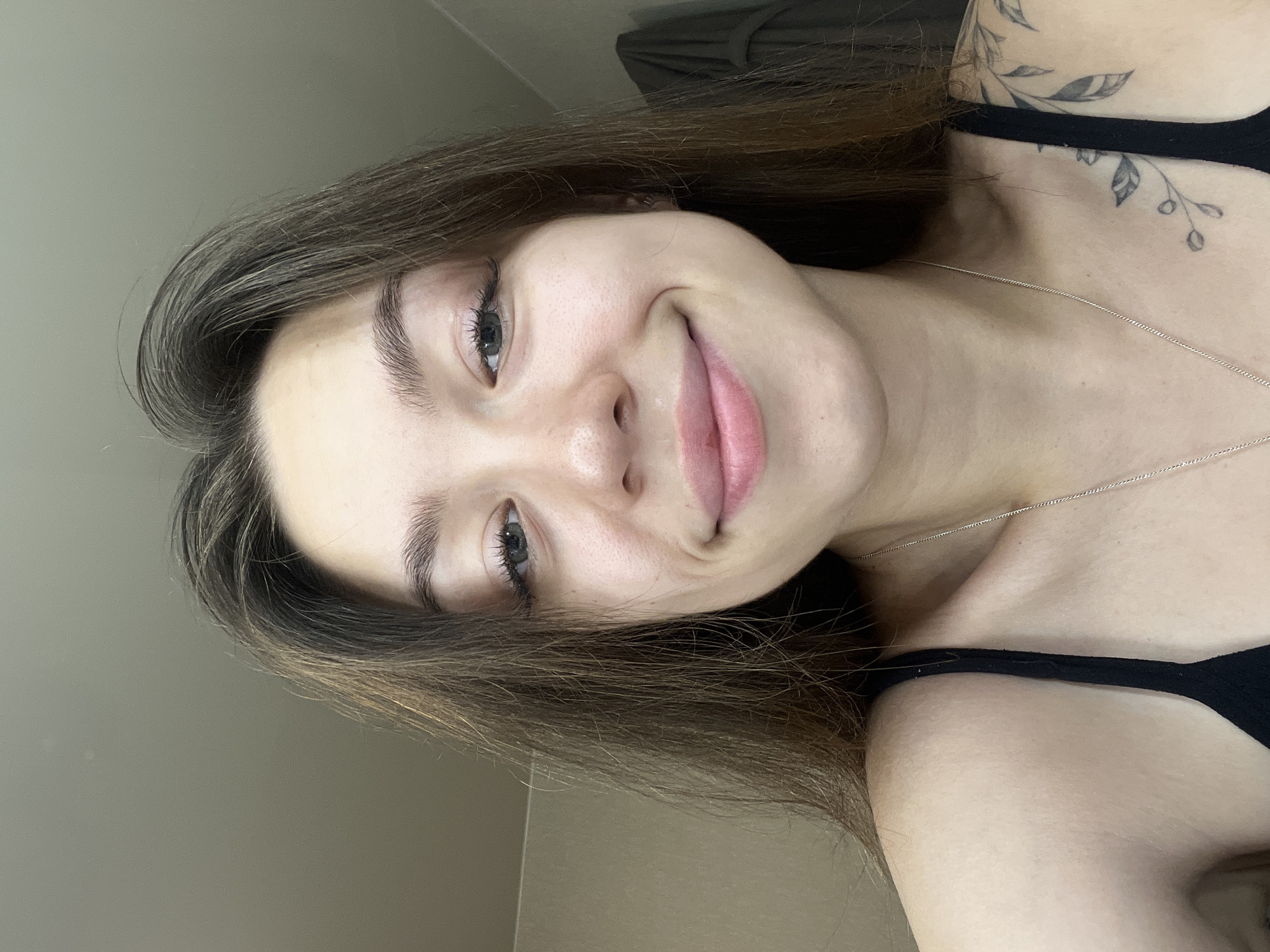 A medium shot captures a young woman with long, brown hair, looking directly at the camera. She is wearing a black tank top and a thin silver necklace. Her skin appears clear with light makeup, and she has a gentle smile. The background is a plain, light-colored wall. The image is a selfie, taken indoors.