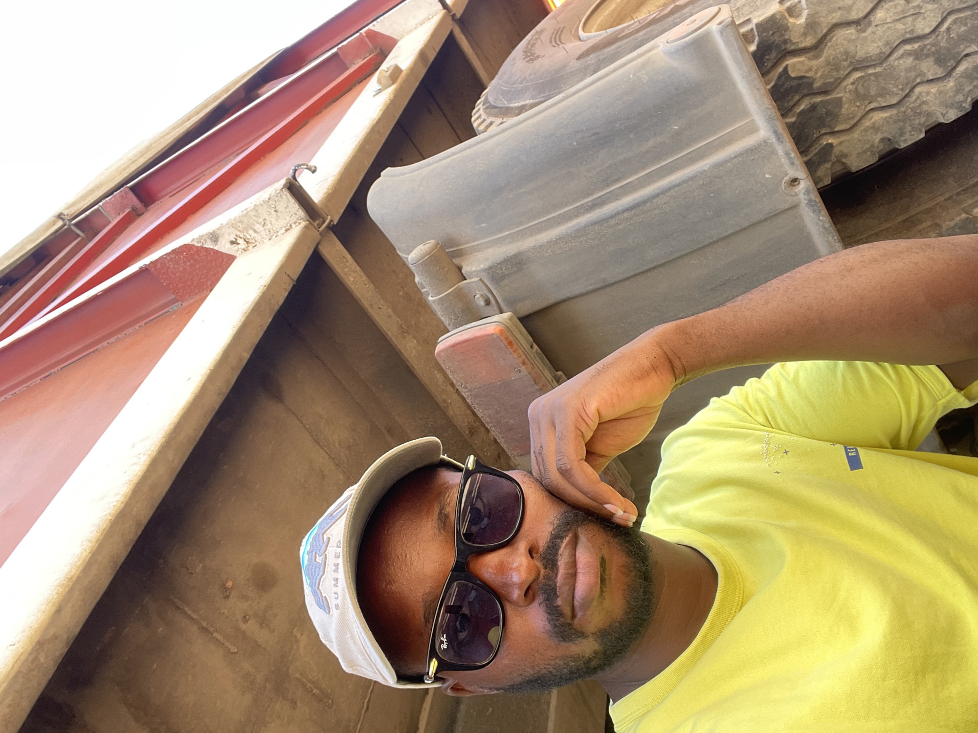 A man wearing a light yellow shirt, sunglasses, and a cap with "SUMMER" printed on it is shown in a low-angle shot inside a large vehicle, possibly a truck bed. The interior is dusty and partly red in color. The man has a beard and is holding his hand up to his face. A large tire and a gray component are visible in the background. The image was taken in Abuja, Nigeria.