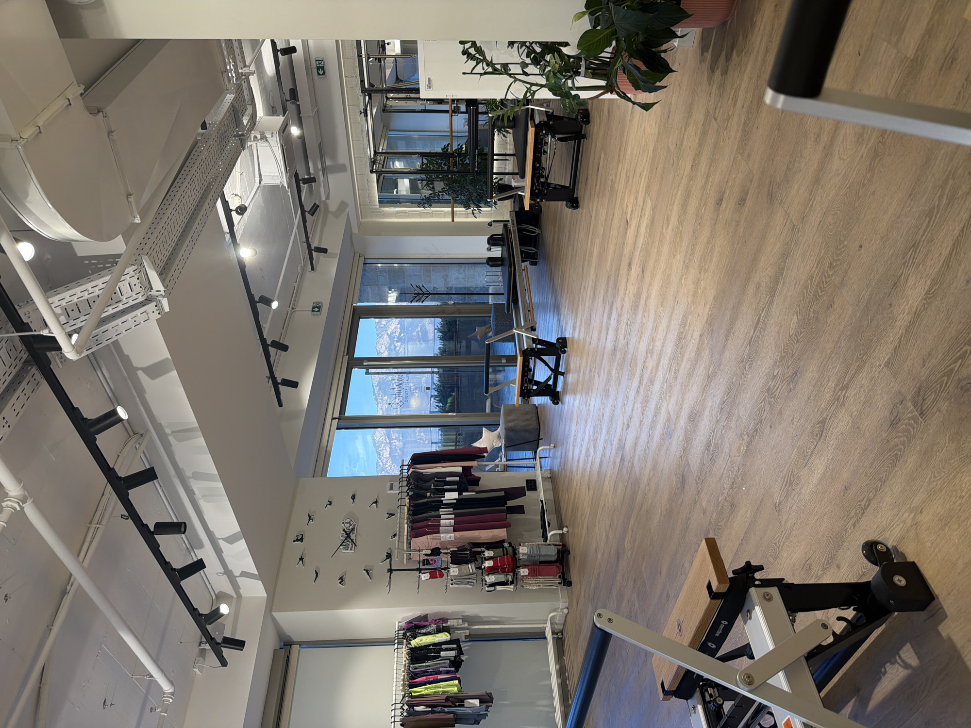 The image depicts the interior of a Pilates studio in Boreti, Montenegro. The studio features a light-colored wooden floor and a high ceiling with exposed industrial ductwork and track lighting. On the left side of the room, there are clothing racks displaying athletic wear, and a wall-mounted clock with a unique design. Several Pilates reformers, characterized by their wooden and metal frames with black padding, are positioned throughout the space. Large windows with a mountain view in the background are visible. Potted plants are placed in strategic locations, adding a touch of greenery. The overall impression is a clean, modern, and well-lit fitness studio. There are no people visible in the image.