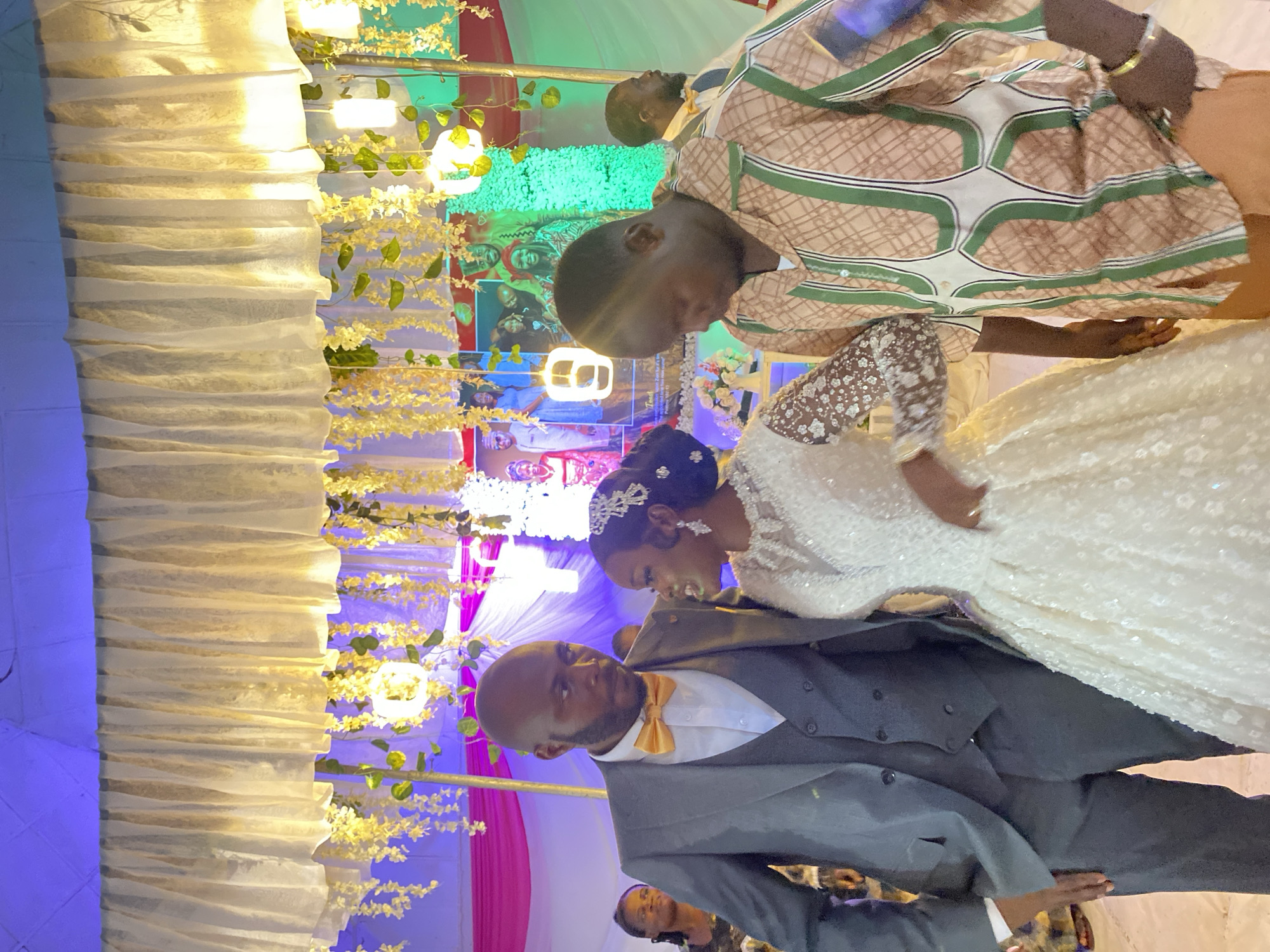 The image captures a wedding reception in Vom, Nigeria. The bride and groom, dressed in a white gown and a grey suit with a yellow bow tie, respectively, are the focal point. They are standing close to each other, seemingly interacting.
Behind them is a decorated backdrop featuring a green and pink design, with images of the couple displayed. Wedding decorations, including draped white fabric and strings of yellow flowers, are visible on the left. To the right and behind the couple is a male wearing a patterned brown and white shirt. The environment is illuminated with artificial lighting.