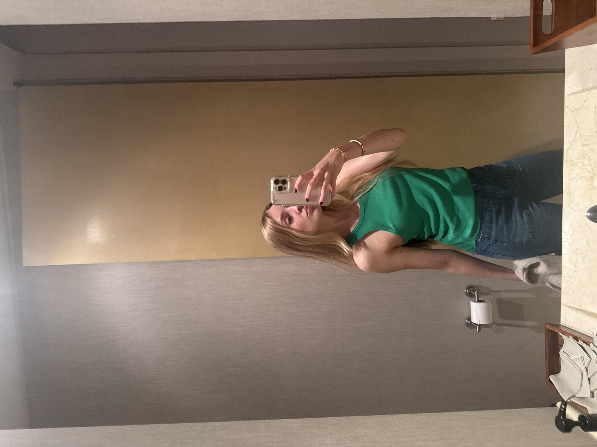 A woman is taking a mirror selfie in a bathroom. She is wearing a green tank top and blue jeans. The bathroom has a light brown upper wall and a gray lower wall. There is a toilet paper holder on the wall and a shelf with towels. The location is Mexico City, Mexico.