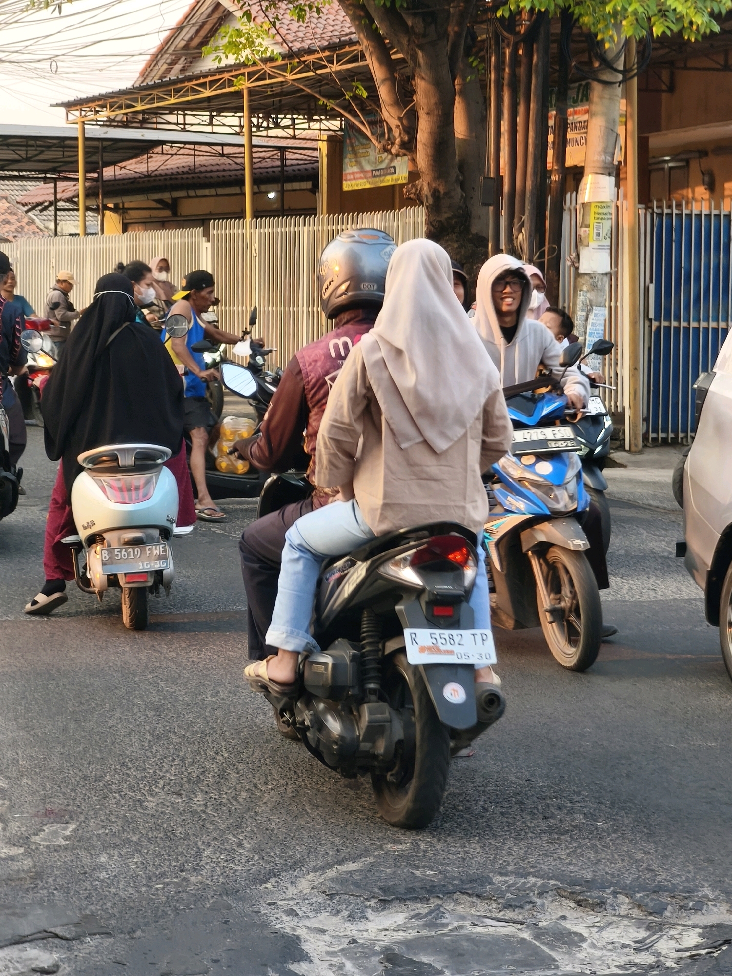 The image depicts a street scene in Satriamekar, Indonesia, with several people riding motorcycles. In the foreground, a person wearing a light brown hijab and blue jeans rides on the back of a motorcycle, with their legs visible. The motorcycle has a license plate that reads "R 5582 TP 05-30". To the left of this motorcycle is another lighter-colored scooter with a license plate "B 5619 FHE 10-36". On this scooter, a person in a black abaya and red pants, with a head covering and a surgical mask, is seated. Further to the left, a person in a blue shirt and dark shorts rides a motorcycle. In the midground, a motorcycle with a blue fairing is driven by a person wearing a grey hoodie and glasses. Other individuals and motorcycles are partially visible in the background. The environment includes a building with a tiled roof and yellow structural elements, a white slatted fence, and trees. The road surface is asphalt with visible cracks and patches. The lighting suggests it is late afternoon or early evening.