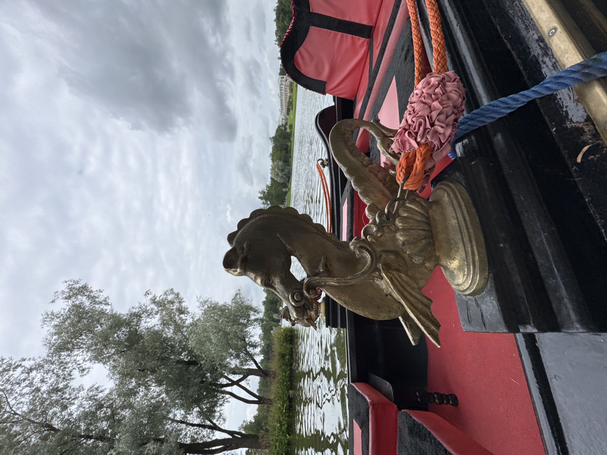 The image captures a view from a gondola-style boat on a canal in the Catherine Park, city of Pushkin, Russia. In the foreground, a golden statue of a mythical sea creature adorns the side of the boat. The boat has red and black seating with orange and blue ropes. The canal water is calm with reflections of the trees lining the bank. In the background, a grand white building with classical architecture is visible across the water, characteristic of the park's palaces. The sky is overcast with grey clouds, suggesting a cool, humid day. There are no people visible in the image.