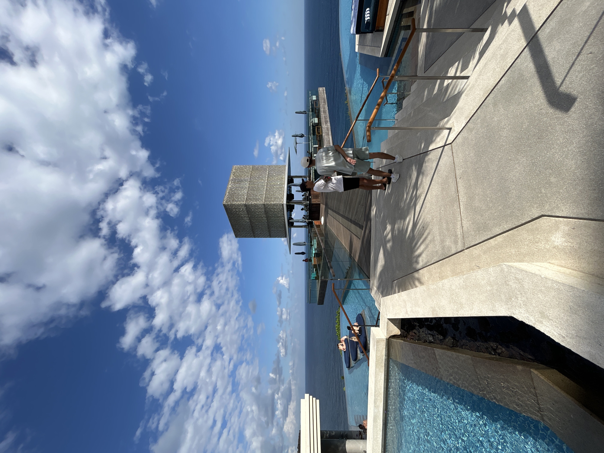 The image shows a resort in Kuta Selatan, Indonesia. Two men stand on a walkway looking out at the ocean. In the foreground, a swimming pool is visible. In the background, an infinity pool overlooks the ocean. There are lounge chairs and umbrellas set up around the pool. The sky is blue with some clouds. The architecture of the resort is modern and sleek. The overall impression is one of luxury and relaxation.
