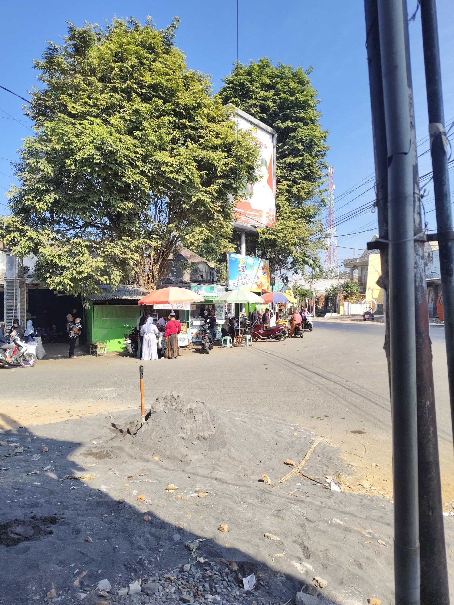 The image depicts a street scene in Gambirono, Indonesia. In the foreground, a pile of dark sand and gravel with a shovel is present, casting a shadow on the ground. The middle ground features several food stalls with colorful umbrellas and awnings, where people are gathered. Some individuals are standing, conversing, and tending to the stalls, while others are seated or interacting with motorcycles. Motorbikes are parked and in use, suggesting local transportation. Behind the stalls, large trees provide shade, and buildings line the street. In the background, a tall telecommunications tower is visible against a clear blue sky. Electrical wires are strung across the scene, indicating infrastructure. The overall impression is of a bustling local marketplace or roadside food area.