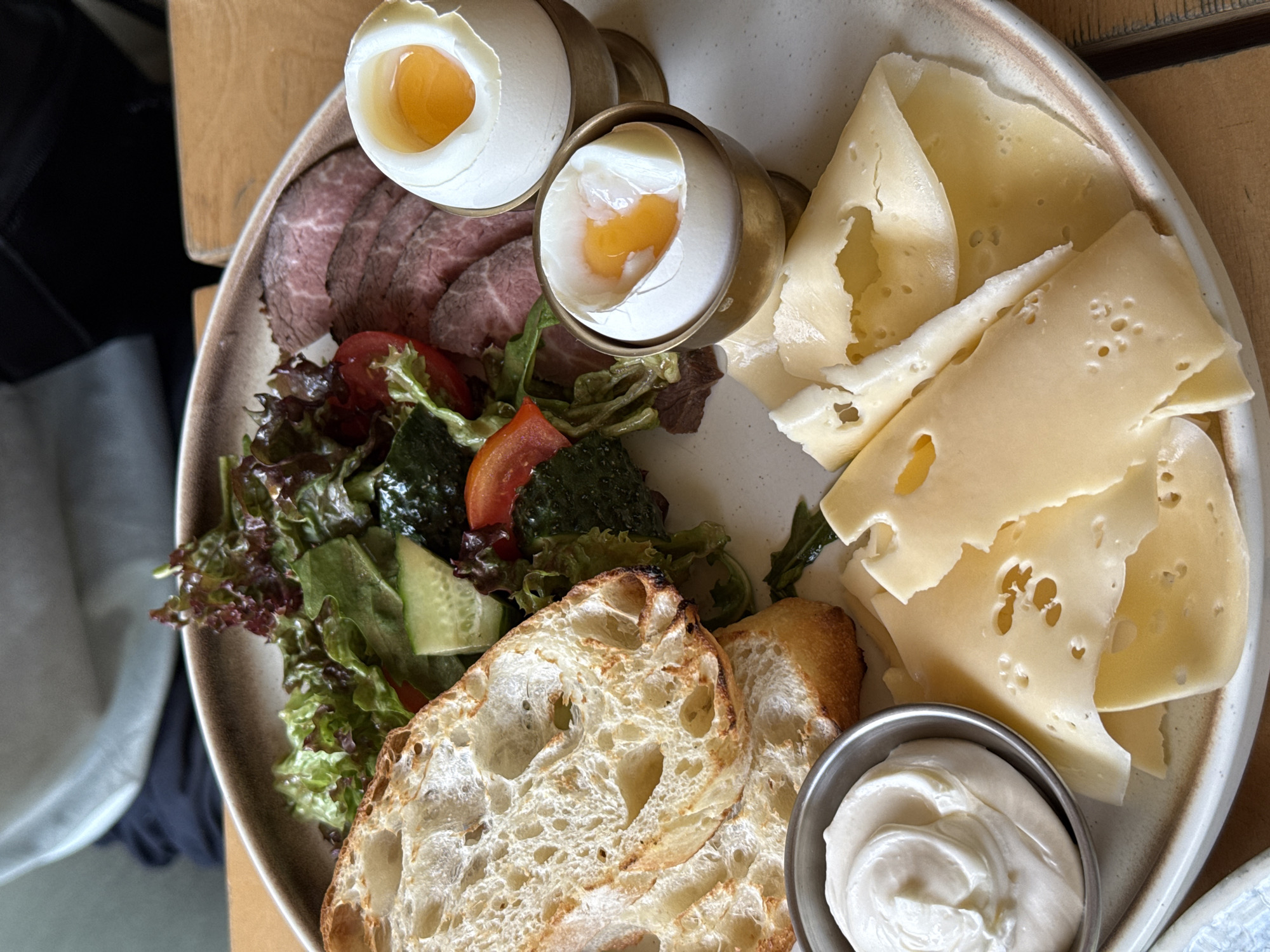 A close-up, high-angle shot showcases a breakfast or brunch platter on a wooden table. The platter, a light-colored ceramic dish with a brown rim, is filled with various food items.
In the upper left are two slices of crusty bread with an open crumb structure and toasted surfaces. Adjacent to the bread is a salad, featuring red leaf lettuce, cucumber slices, and red tomato wedges, with some darker green leafy vegetables also visible.
To the right of the salad are several thin slices of cooked roast beef, appearing to be medium-rare. Two soft-boiled eggs in brass egg cups are positioned to the right of the roast beef. The top egg is slightly more cooked, with a more solidified yolk.
In the bottom left of the platter is a small metal bowl filled with a creamy white substance, possibly cream cheese or sour cream, with visible swirls on its surface. To the right of the bowl and extending towards the bottom edge of the image are several thin slices of cheese with distinct holes, resembling Swiss cheese.
The background is blurred, but hints of a person's shoulder wearing a light-colored garment and dark fabric suggest the presence of a diner. The overall lighting is natural and bright, suggesting an indoor setting with good illumination.