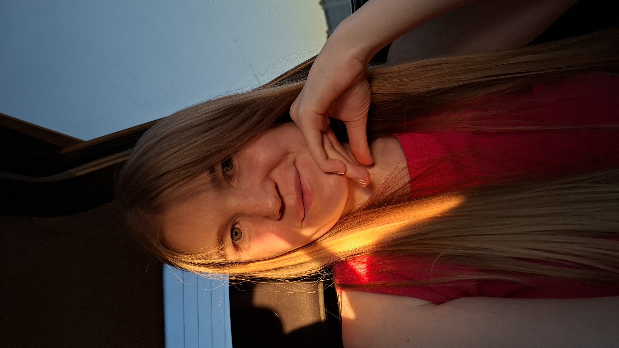 A young, fair-skinned woman with long, straight, light blonde hair poses in a car. She is wearing a red shirt and her left hand is resting on her chin. She is smiling slightly at the camera. The car sunroof is open, revealing a clear sky at dusk. Light from the setting sun illuminates her face and hair. The location is Chaikovsky, Russia.