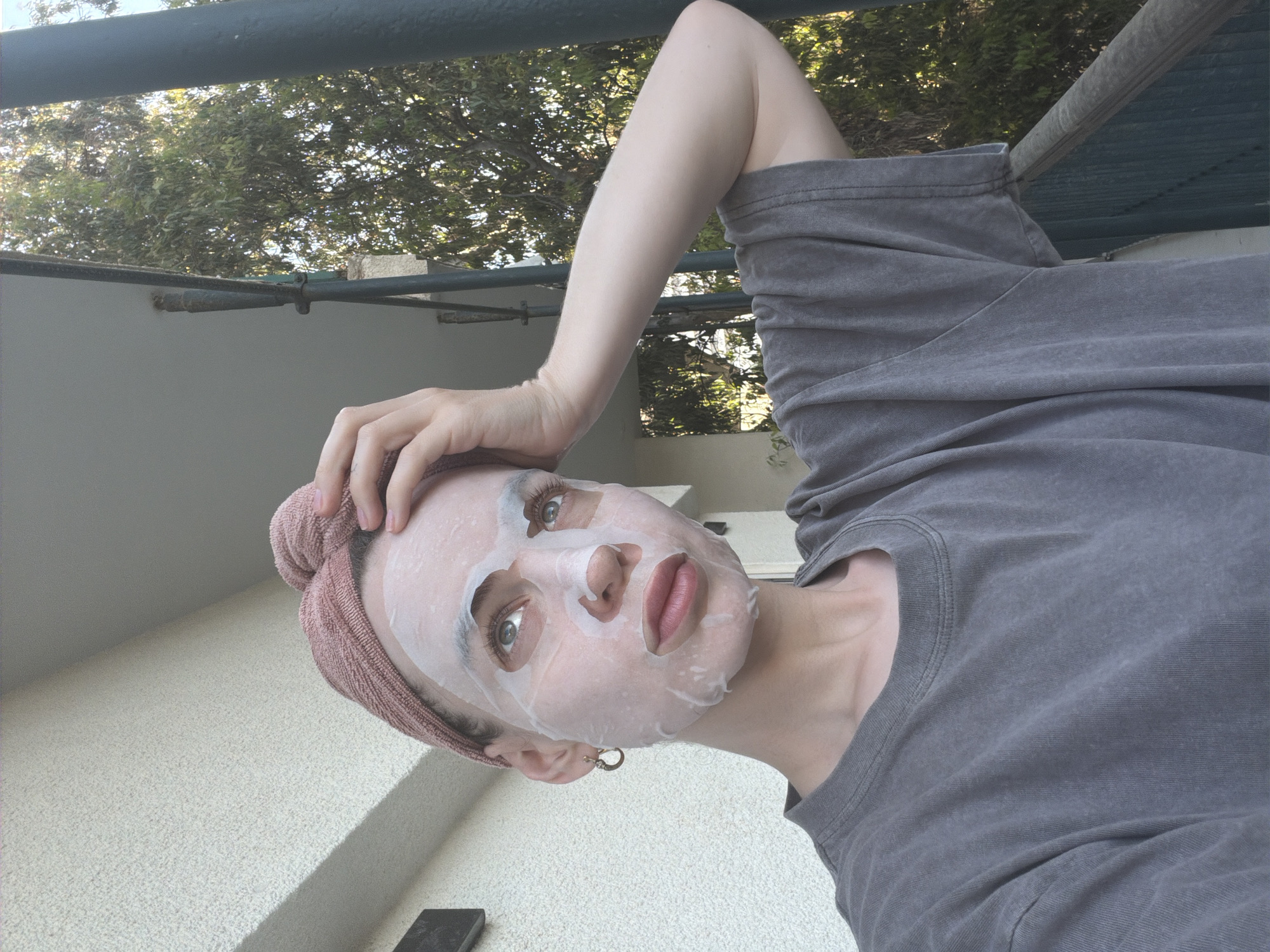 A woman with a sheet mask on her face is posing for a photo outdoors. She's wearing a gray t-shirt and has a pink towel wrapped around her head. Her arm is raised above her head, and her face is tilted towards the camera. The background includes a wall with a textured surface, trees, and a metal railing. The location cannot be determined from the provided image.