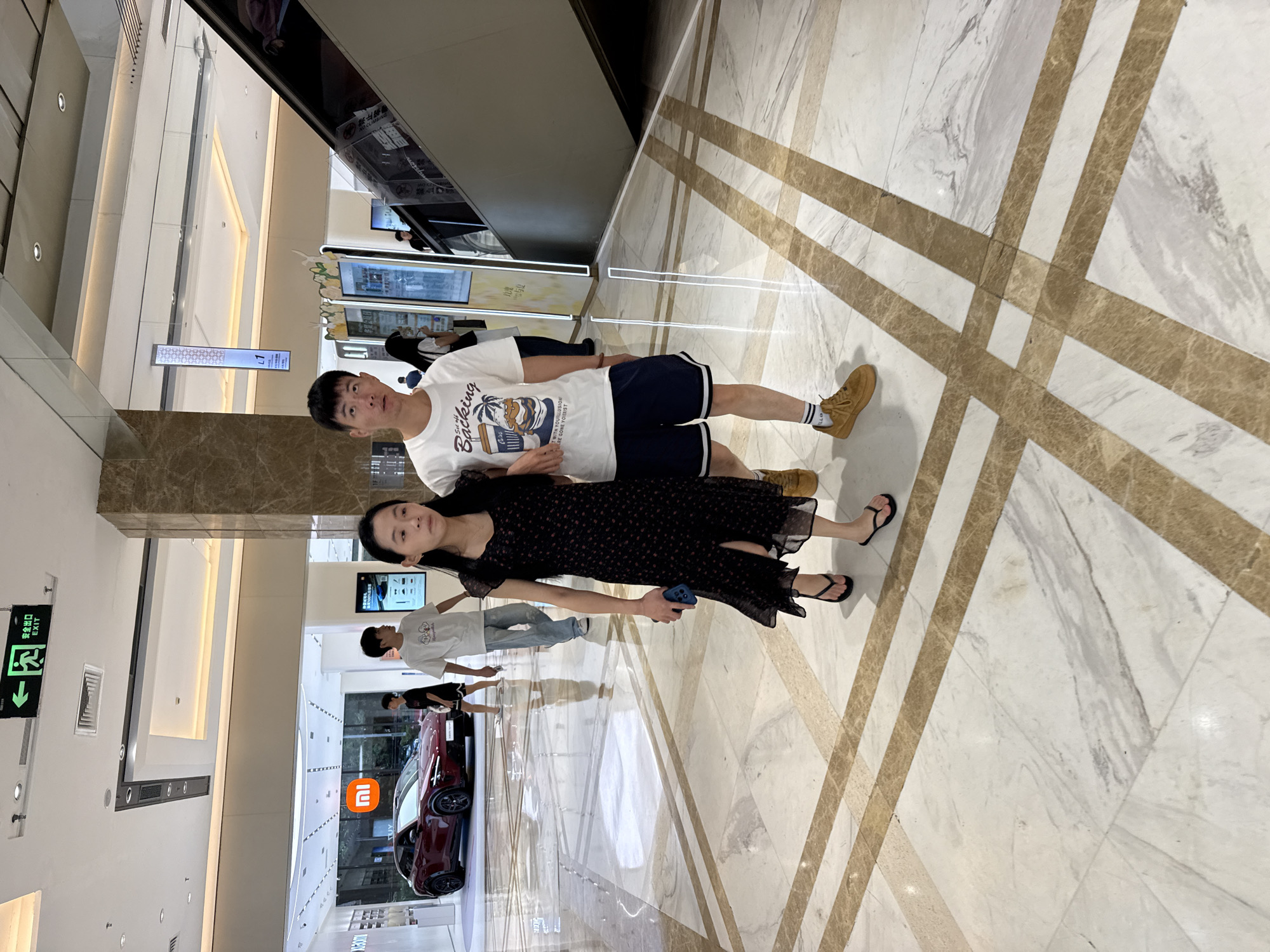 In Huizhou, China, a young couple is walking on a polished marble floor in a brightly lit indoor commercial space. The man, wearing a white t-shirt with a graphic print and navy blue shorts, is on the right. The woman, in a black floral print dress and black flip-flops, is on the left, with her left arm around the man's waist. They are walking away from the camera, towards the left side of the frame, in front of a row of display screens and an escalator. In the background, a red electric car is displayed in a retail showroom. An "EXIT" sign with an arrow is visible on the ceiling. The floor features a geometric pattern of inlaid marble.