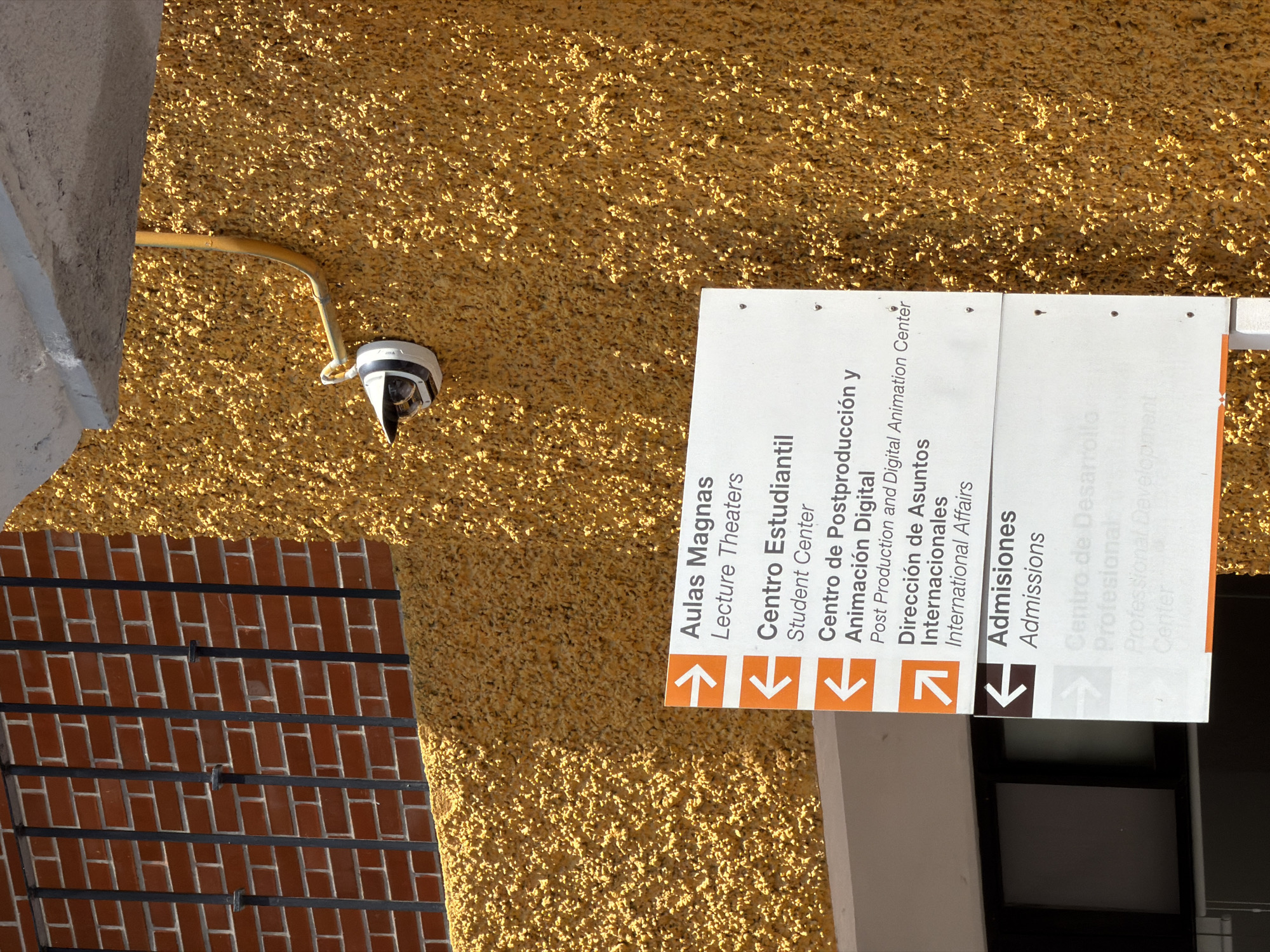 The image shows a building exterior in San Andrés Cholula, Mexico. The building's facade is textured and has a yellowish hue. Attached to the wall is a white directional sign with black text in both Spanish and English, indicating directions to locations such as "Aulas Magnas" (Lecture Theaters), "Centro Estudiantil" (Student Center), "Centro de Postproducción y Animación Digital" (Post Production and Digital Animation Center), "Dirección de Asuntos Internacionales" (International Affairs), and "Admisiones" (Admissions). Arrows point in different directions below each location's name. A security camera is mounted on the wall, positioned to monitor the area. A section of a brick wall with metal support bars is visible on the left side of the frame. A window is barely visible, located behind the sign.