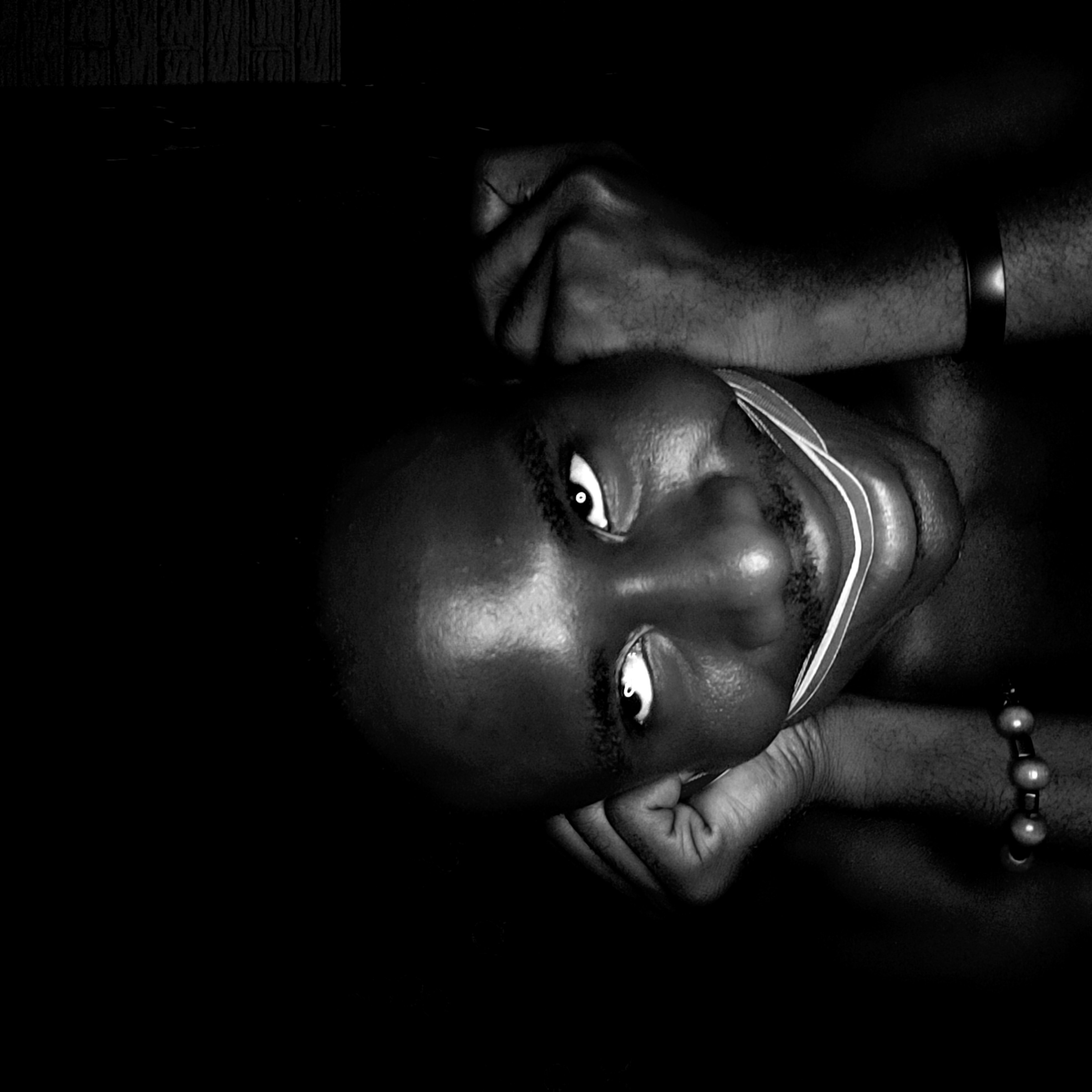 The image is a black and white close-up of a person's face and hands, angled slightly upward. The person appears to be dark-skinned with a shaved head. The eyes are open and looking directly at the viewer. A light-colored band is visible around the neck, possibly a strap or piece of cloth. The person's hands are raised, with the fists close to their face, and a wrist is visible with a bracelet. The background is dark and indistinct. The image is unlikely to be linked to the city of Narayi, Nigeria, as the content of the image doesn't imply that it was taken there.