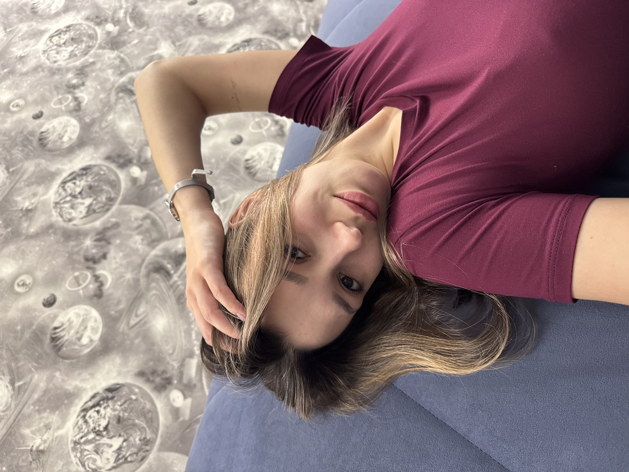 The image shows a woman lying on a blue surface, possibly a couch or bed, in what appears to be an interior setting. She is wearing a maroon-colored short-sleeved shirt and a silver wristwatch. Her hair is blonde and brown and is spread out on the blue surface. She is looking towards the camera with a slight smile. One hand is raised near her head, possibly touching her hair. The background is a light grey surface with a pattern of planets. The location is not discernible from the image.