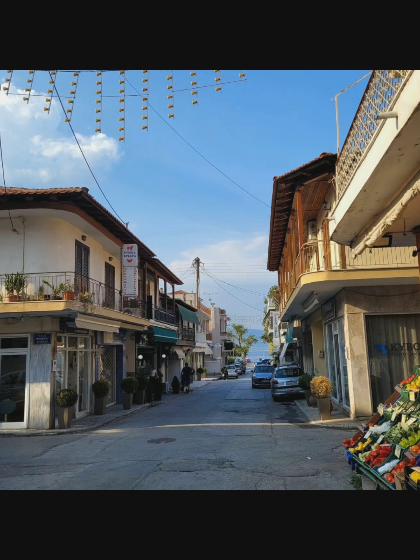 The image depicts a street scene with buildings lining both sides of a paved road. The sky is blue with scattered clouds. String lights are visible overhead, spanning across the street. On the left side of the street, a building with a balcony adorned with potted plants is prominent. A sign with Greek text and a graphic of a pig is affixed to the building. Further down the street, on the right side, a building with a wooden balcony and railing is visible. A fruit stand is set up on the sidewalk in the foreground on the right, displaying a variety of colorful fruits and vegetables. Several cars are parked along the street. A few people are present, walking or standing. The overall impression is of a somewhat quiet street in a European town, possibly a coastal area, given the distant view of water. The location provided, Bielawska Wieś, Poland, is inconsistent with the Greek signage in the image, suggesting a potential error in the location data.