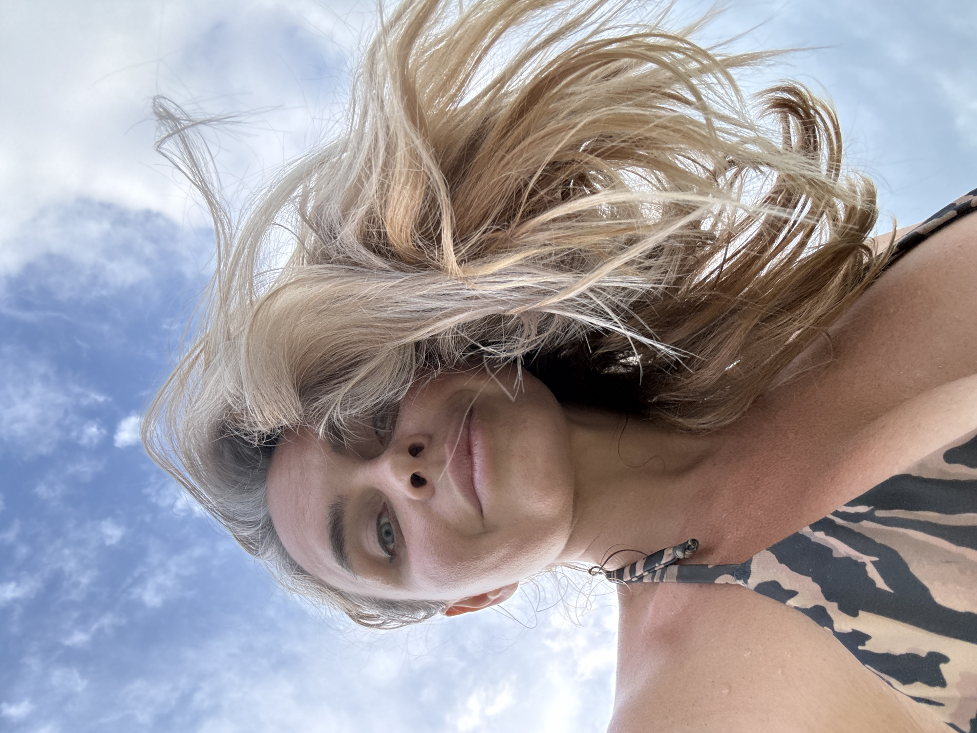 A woman is captured in a low-angle, close-up selfie against a cloudy sky. Her blonde hair is being blown by the wind, framing her face. She is wearing a bikini with a tan and black animal print pattern. The image appears to have been taken outdoors, likely in a tropical location given the lighting and the woman's attire.
