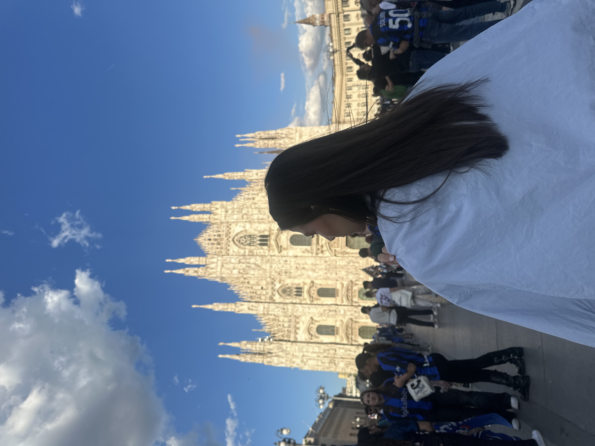 A medium, eye-level, outdoor shot shows a person partially obscuring the camera's view. The person appears to be a young woman with long dark hair and tan skin, with a white garment draped over part of their head and shoulders. The woman is looking down. Behind the woman and to the left is the Duomo di Milano, a large Gothic cathedral with spires, standing under a clear, blue sky with scattered clouds. The cathedral's facade is detailed. Below and in front of the cathedral, a crowd of people can be seen, with many wearing black and blue striped jerseys. Street lamps and architectural details are visible at the bottom of the frame. The image is set in the Piazza del Duomo in Milan, Italy.