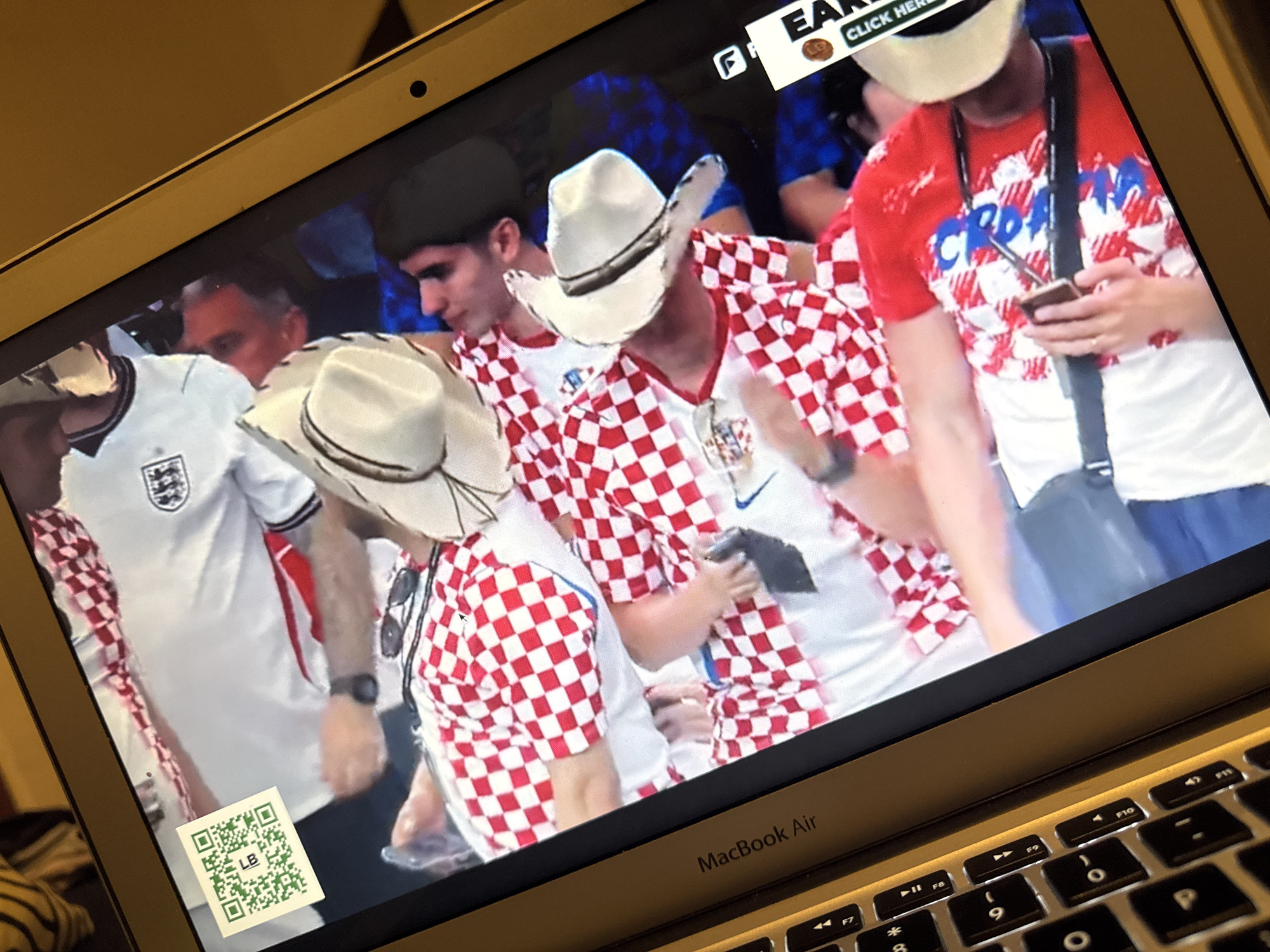 The image displays a laptop screen showing a broadcast of what appears to be a sports event, possibly soccer. Several individuals are visible on the screen, some wearing red and white checkered shirts, which is the national color of Croatia. One person is wearing a white cowboy hat, and another is wearing an England jersey. A QR code with the letters "LB" is affixed to the bottom left corner of the laptop screen. The laptop itself is identified by the "MacBook Air" logo visible on its bezel. The background shows stadium seating and bright lighting typical of a live broadcast. There is no discernible information within the image that places the scene specifically in Kufena, Nigeria.